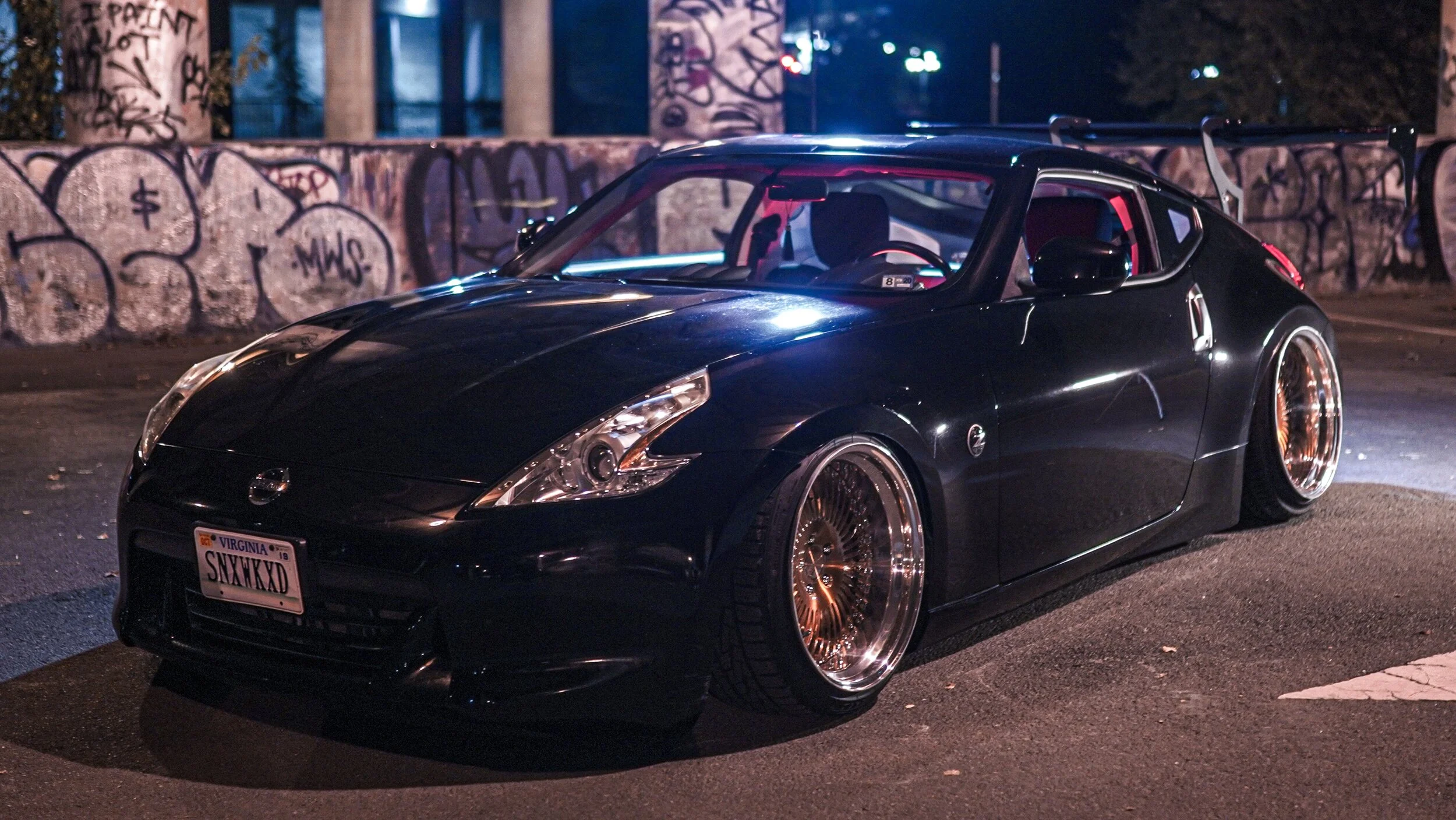 Black Nissan 370Z sports car with custom gold rims parked at night near graffiti-covered concrete pillars.