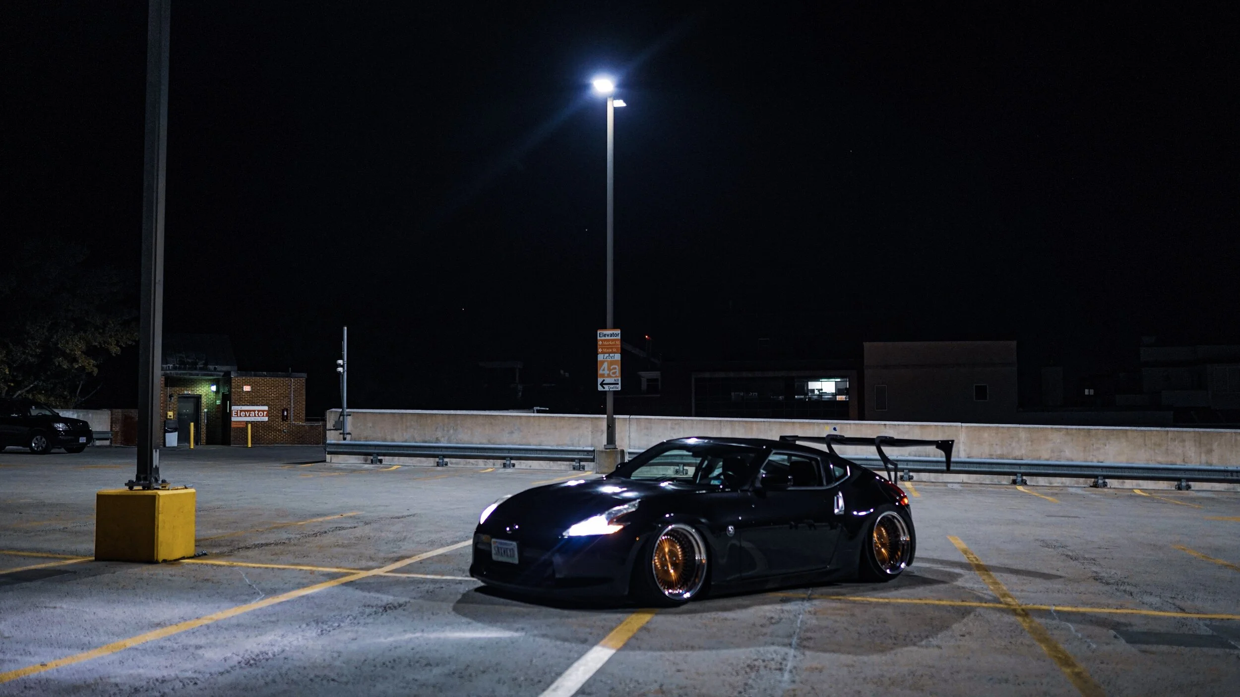 A black sports car with gold rims parked in an almost empty parking lot at night, illuminated by a tall street light.