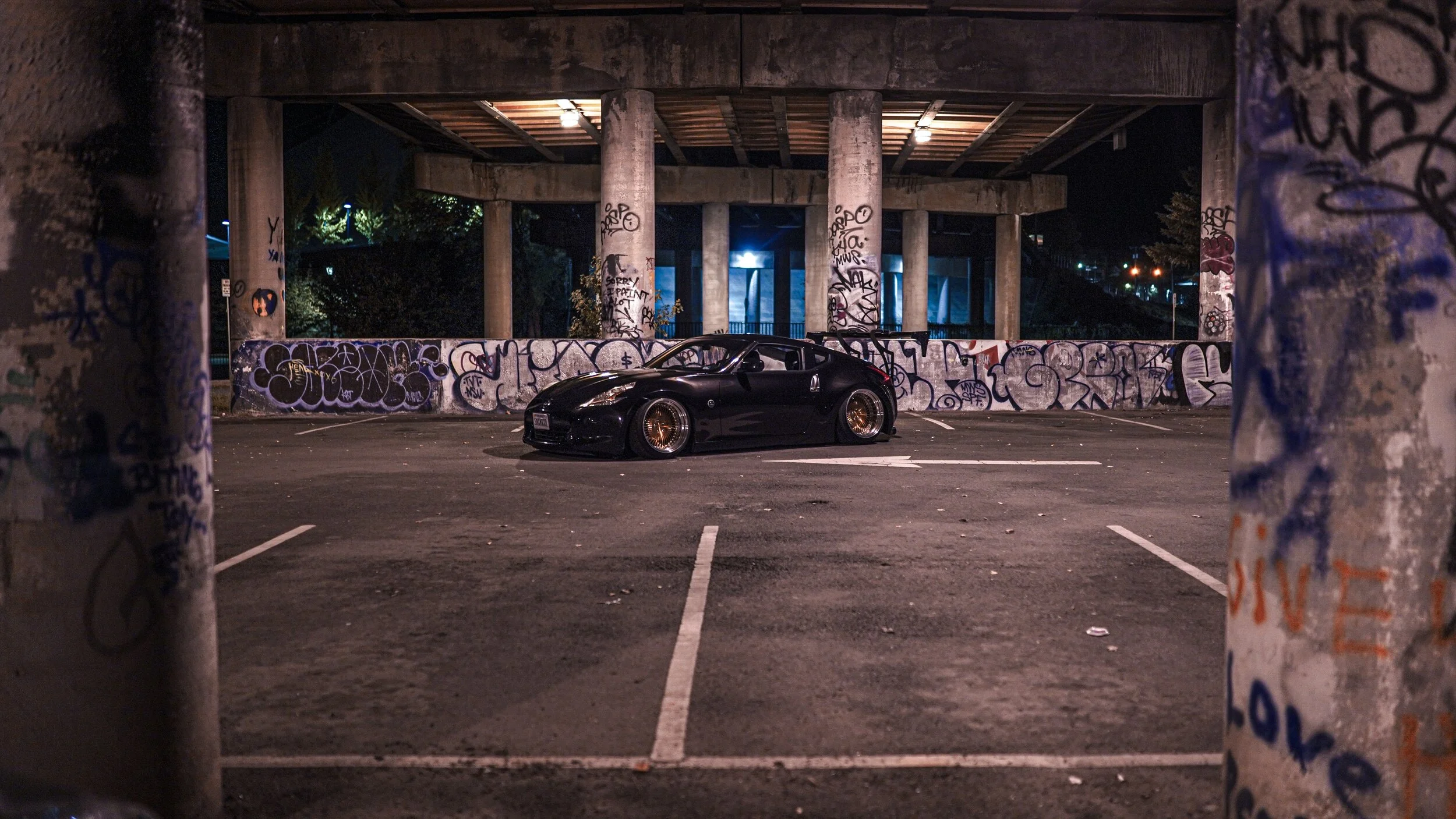 A black sports car with custom bronze wheels parked in an urban lot at night, framed by graffiti-covered pillars and a graffiti wall under an overpass.
