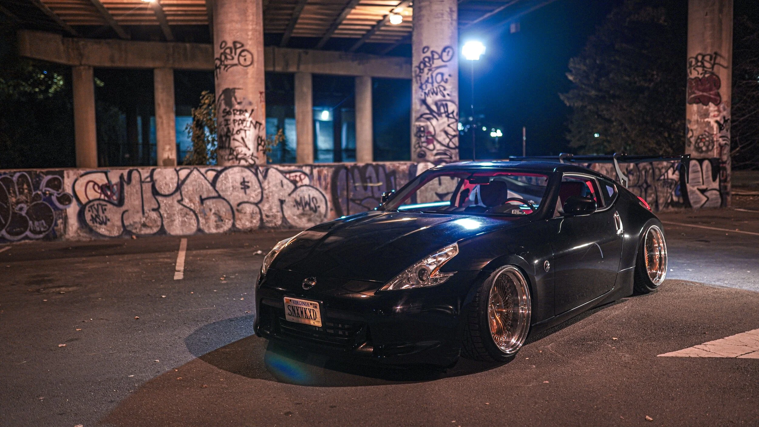A black Nissan sports car with lowered stance, custom wheels, and a large rear wing, parked in an urban area at night with graffiti-covered walls and a concrete support structure in the background.