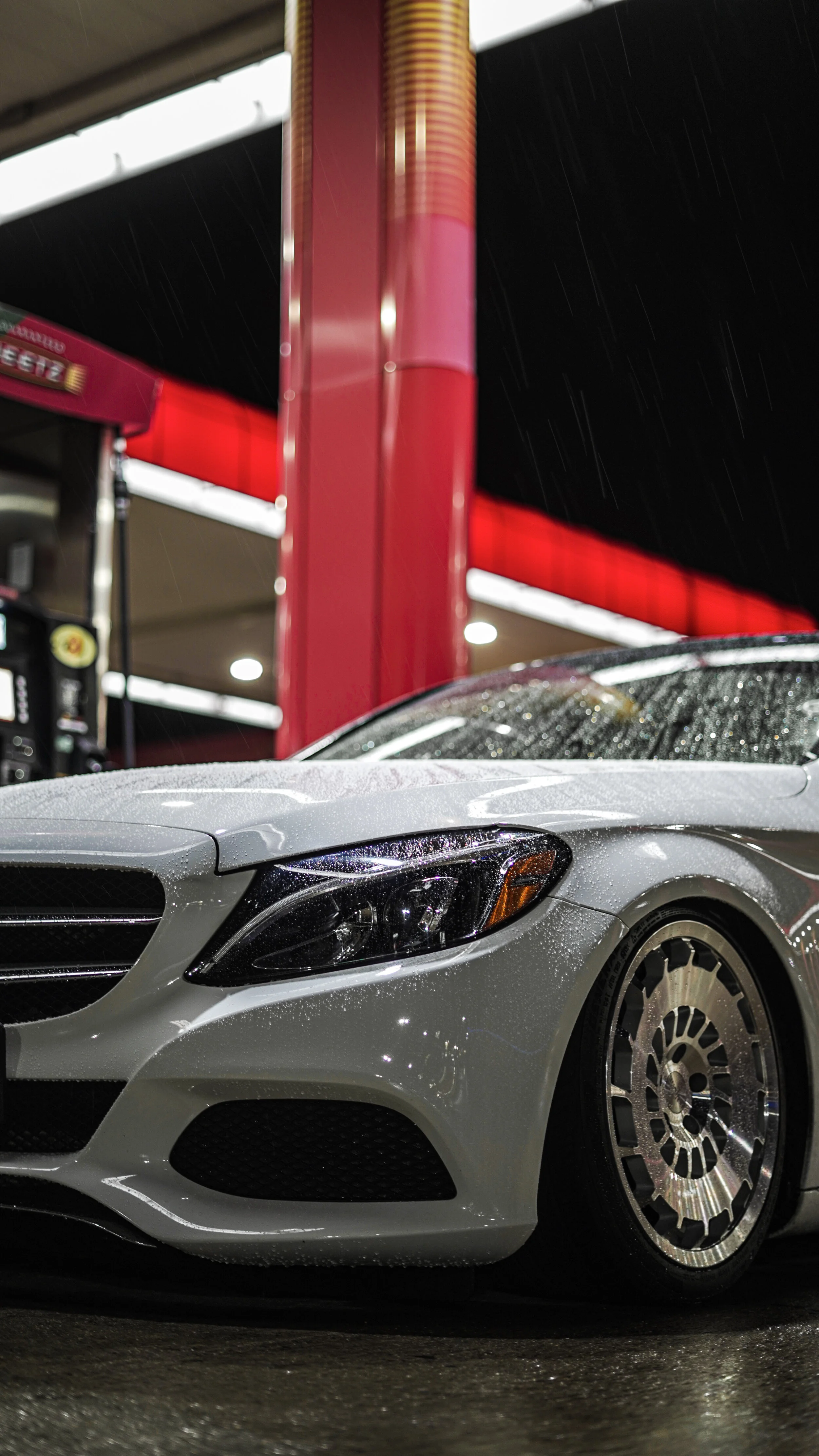 A silver luxury car at a gas station during rain, with water droplets on the car's surface and wet ground.