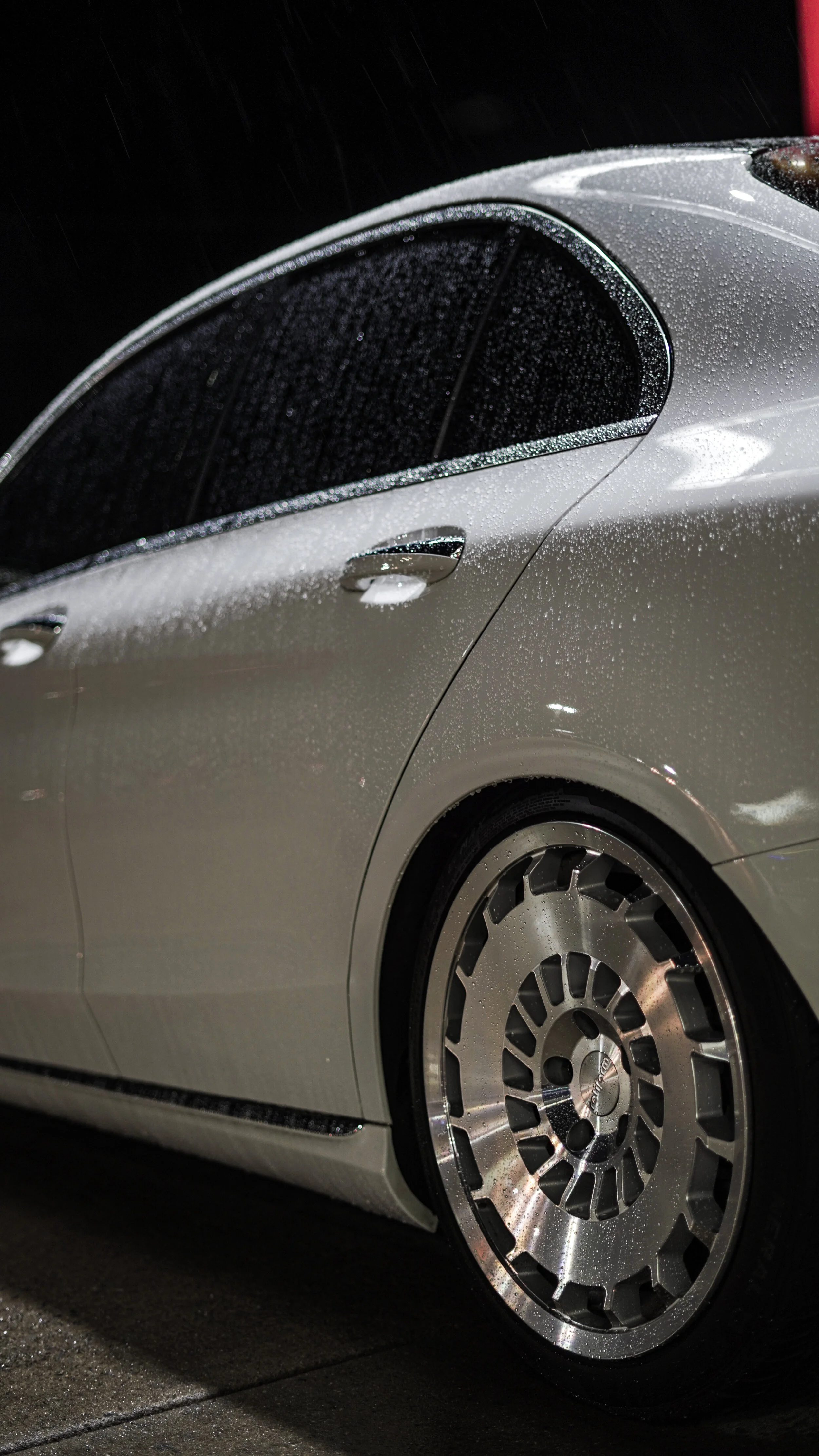 Close-up of a silver luxury car, wet from rain, at night, showing the side panel, rear wheel, and part of the rear window.