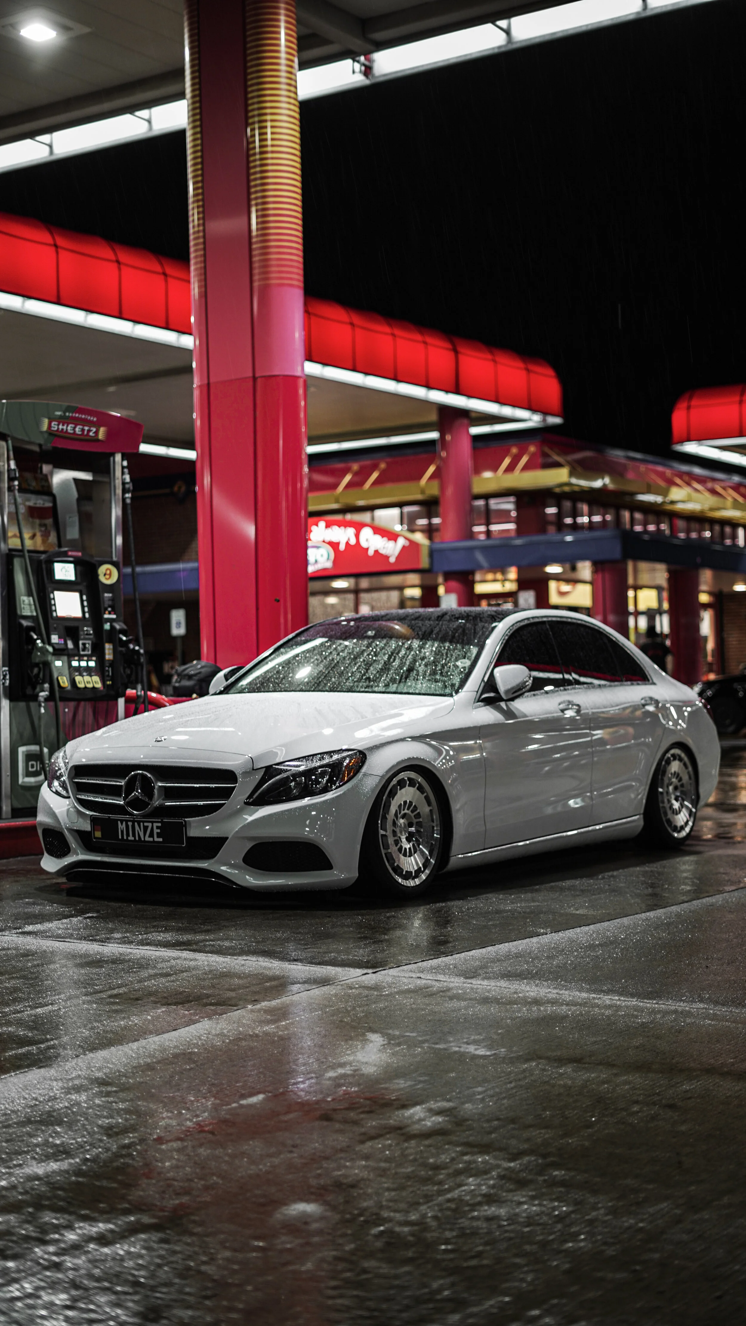A white Mercedes-Benz sedan with custom license plate 'MINZE' parked at a gas station during rain at night. The station has red and black accents with wet pavement reflecting lights.