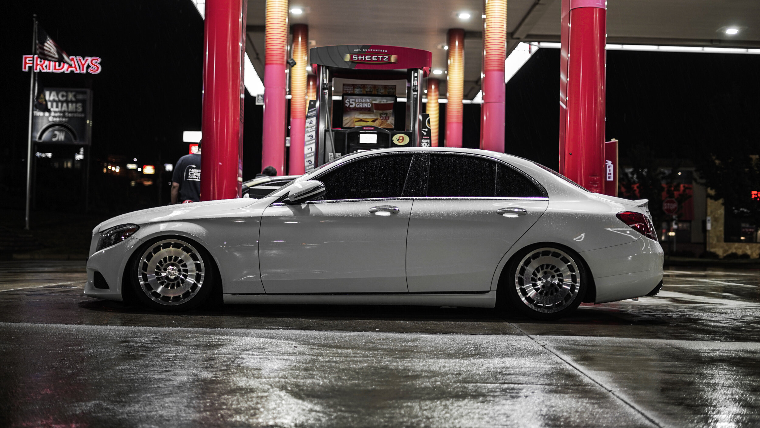 A silver luxury sedan with custom wheels parked at a gas station at night, with wet pavement and illuminated signs in the background.