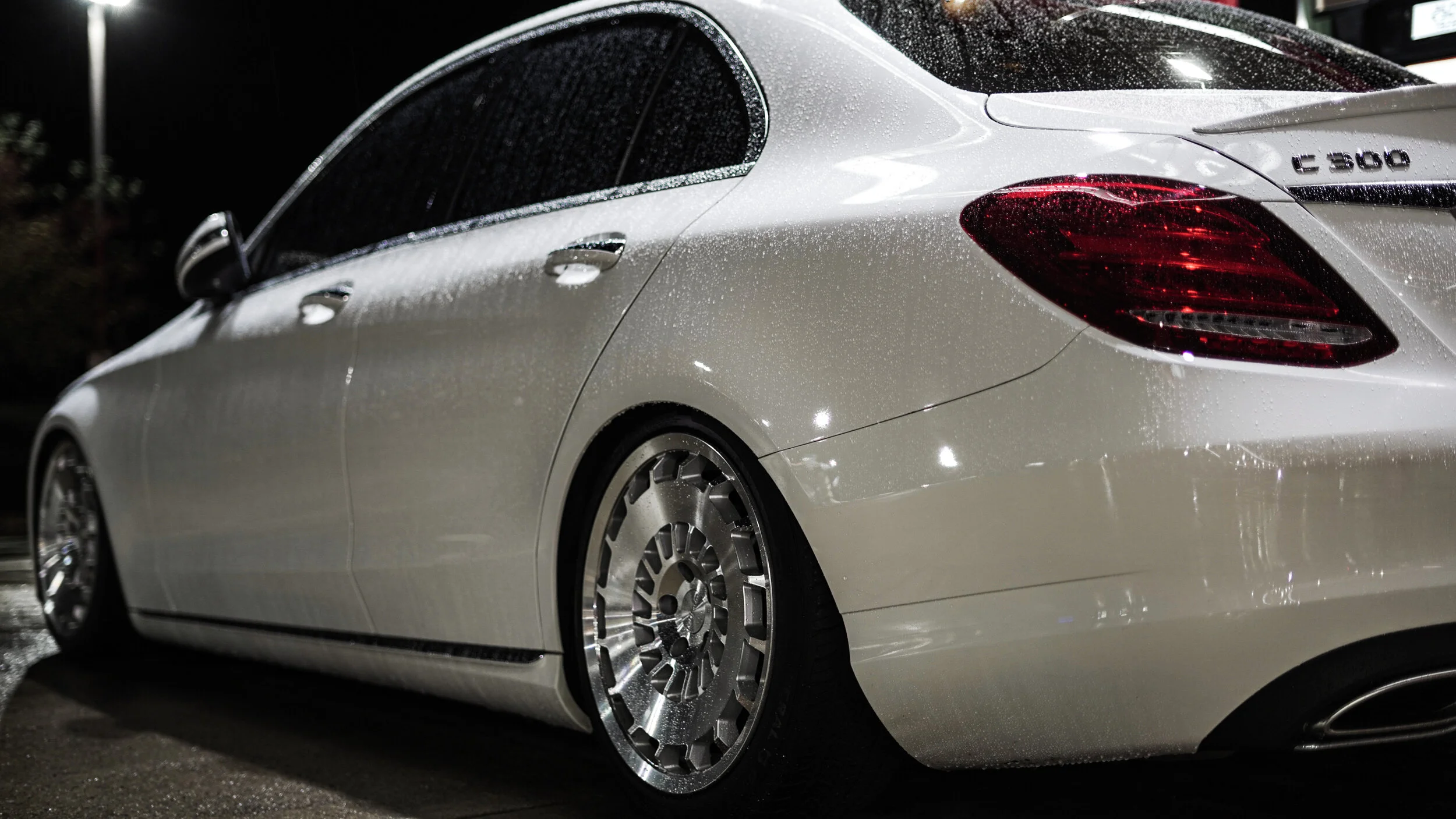 Close-up of a white luxury car, wet from rain, parked at night with reflections of city lights on its surface.