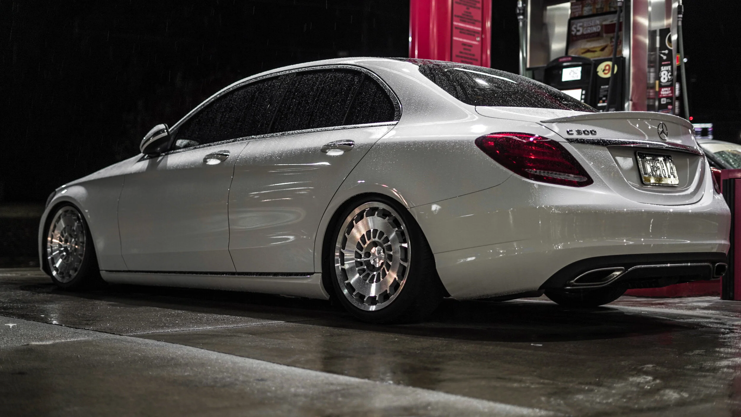 A silver Mercedes-Benz C 300 parked at a gas station at night, wet from rain.
