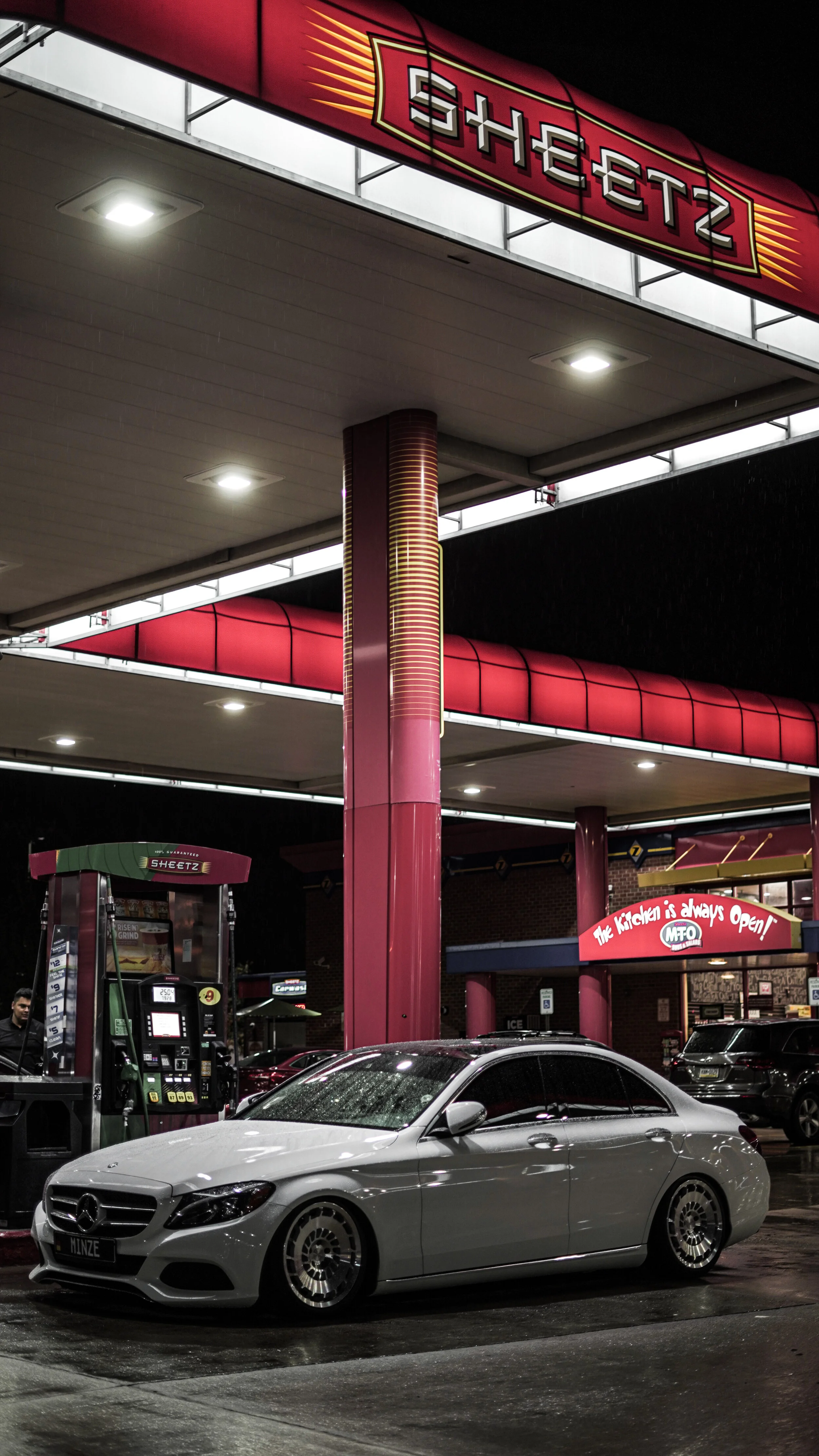 A silver Mercedes-Benz car parked at a Shell gas station at night, with gas pumps and a lit Shell sign overhead.