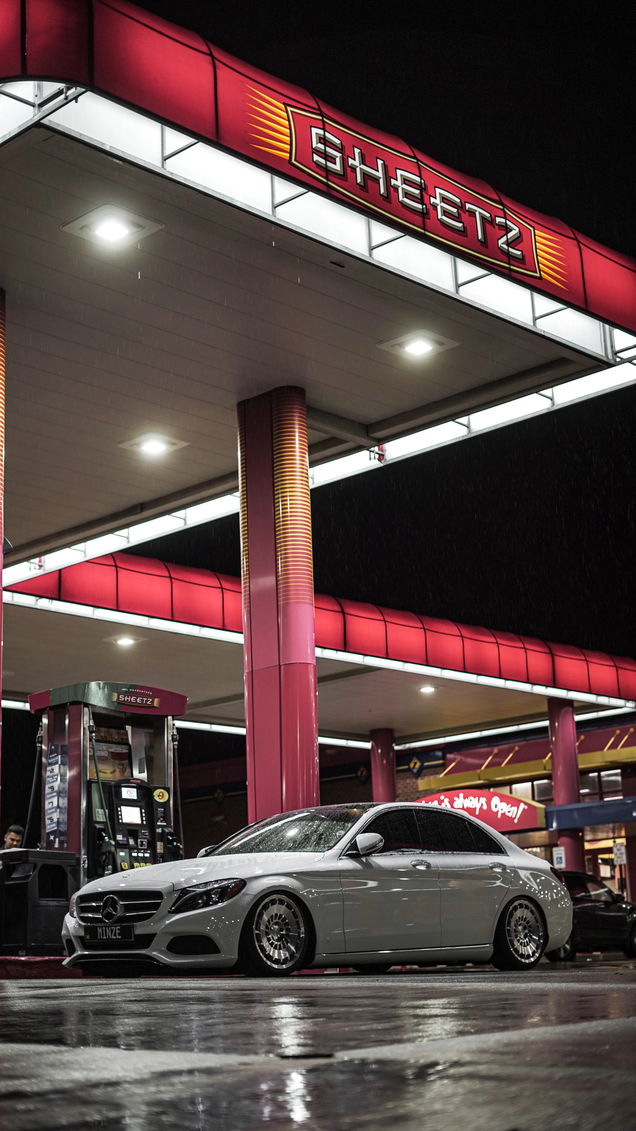 A silver Mercedes-Benz parked at a Sheetz gas station at night, with wet pavement under rain, illuminated sign, and gas pump.
