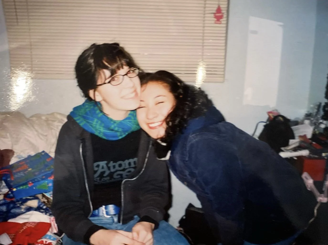 A young man with glasses and a woman with curly black hair smiling and hugging in a cluttered room with presents and a Christmas tree.