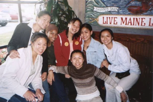 Seven women smiling and posing together indoors, some sitting and some standing, with a mural and window in the background.