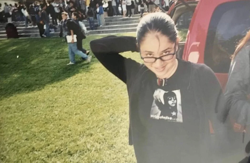 Young woman with glasses and a choker adjusting her hair outdoors, with a crowd and steps in the background.