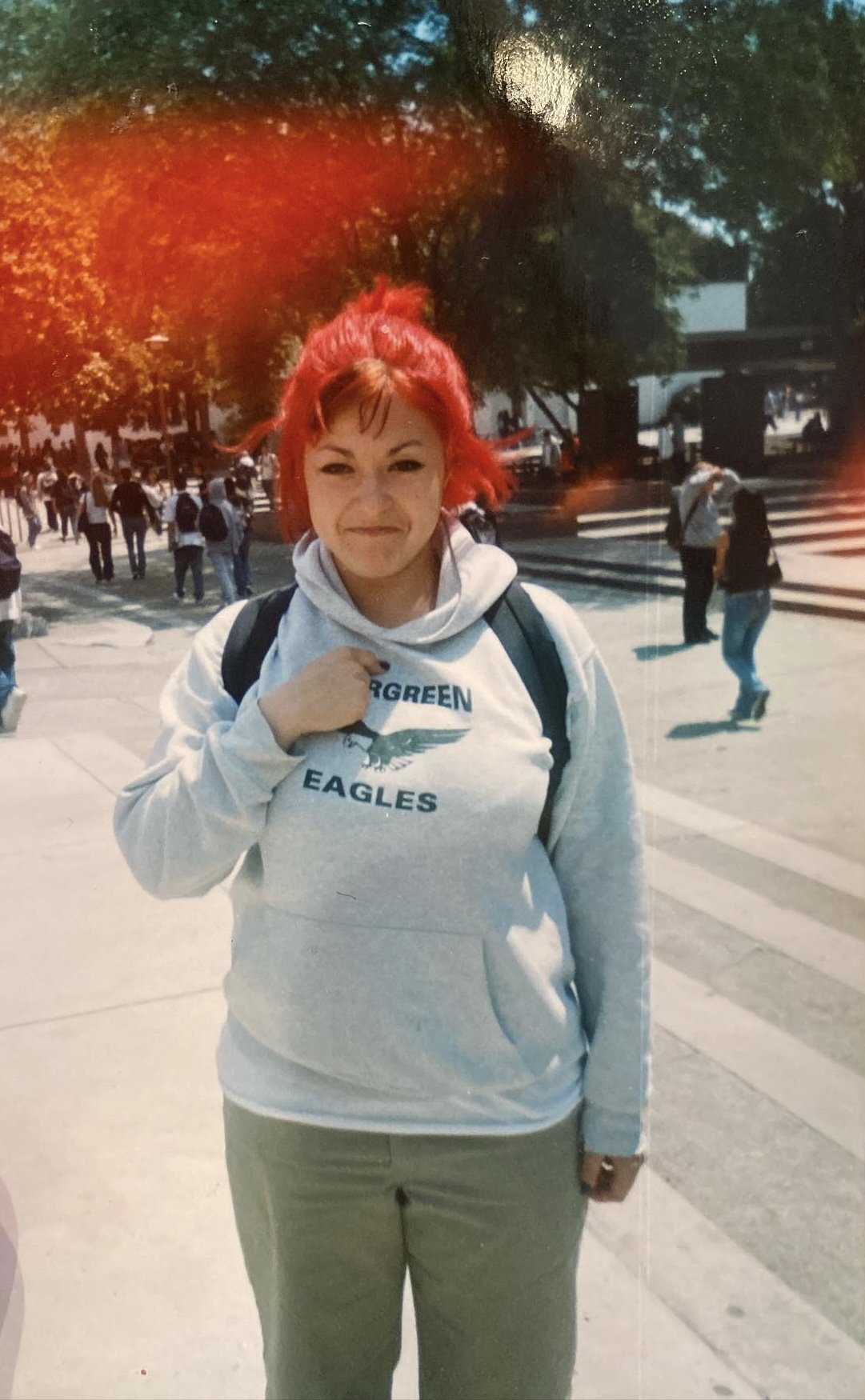 A woman with red hair standing on a city street, wearing a gray hoodie with an eagle logo and the words 'Green Eagles,' carrying a backpack, with other pedestrians and trees in the background.
