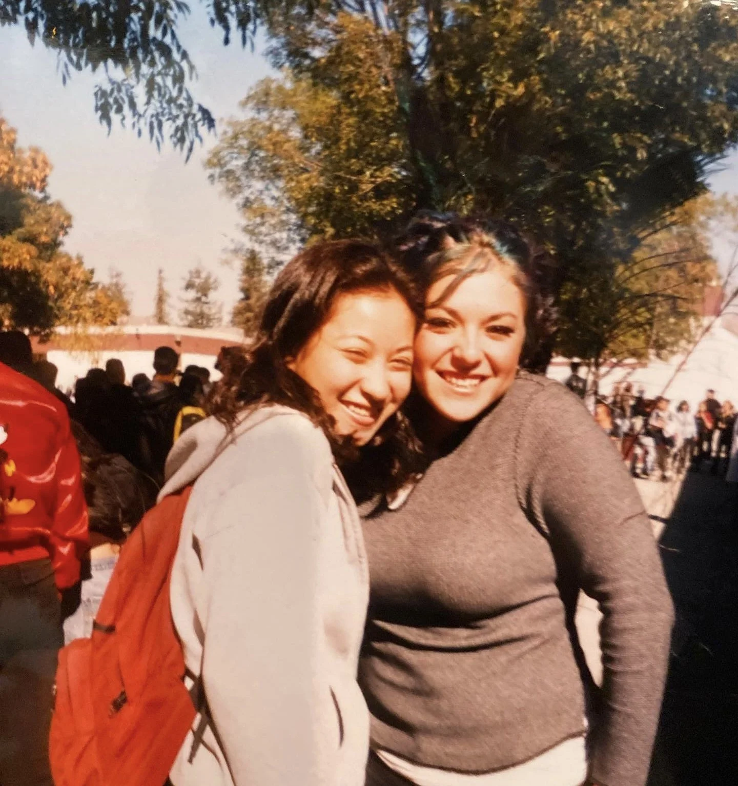 Two young women smiling and hugging each other outdoors during daytime, with trees and a crowd in the background.