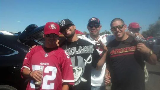Four men standing outdoors in bright sunlight, some wearing San Francisco 49ers jerseys, with a car in the background.