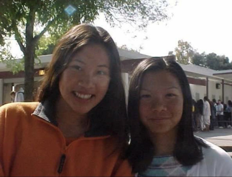 Two women smiling outdoors with a tree and a building in the background.
