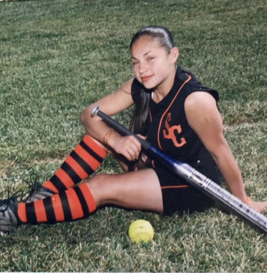 A young girl sitting on grass with a lacrosse stick and a yellow lacrosse ball. She is wearing a black sports outfit with orange details, including striped orange and black socks, and is smiling.