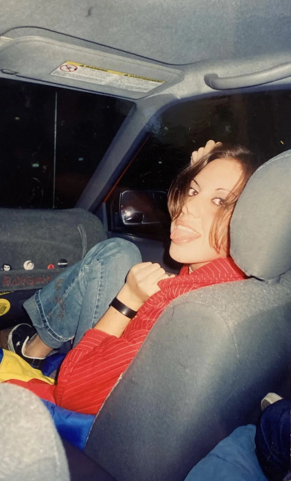 A young woman with shoulder-length brown hair making a face and sticking her tongue out while sitting in the backseat of a car at night, wearing a red jacket and jeans.
