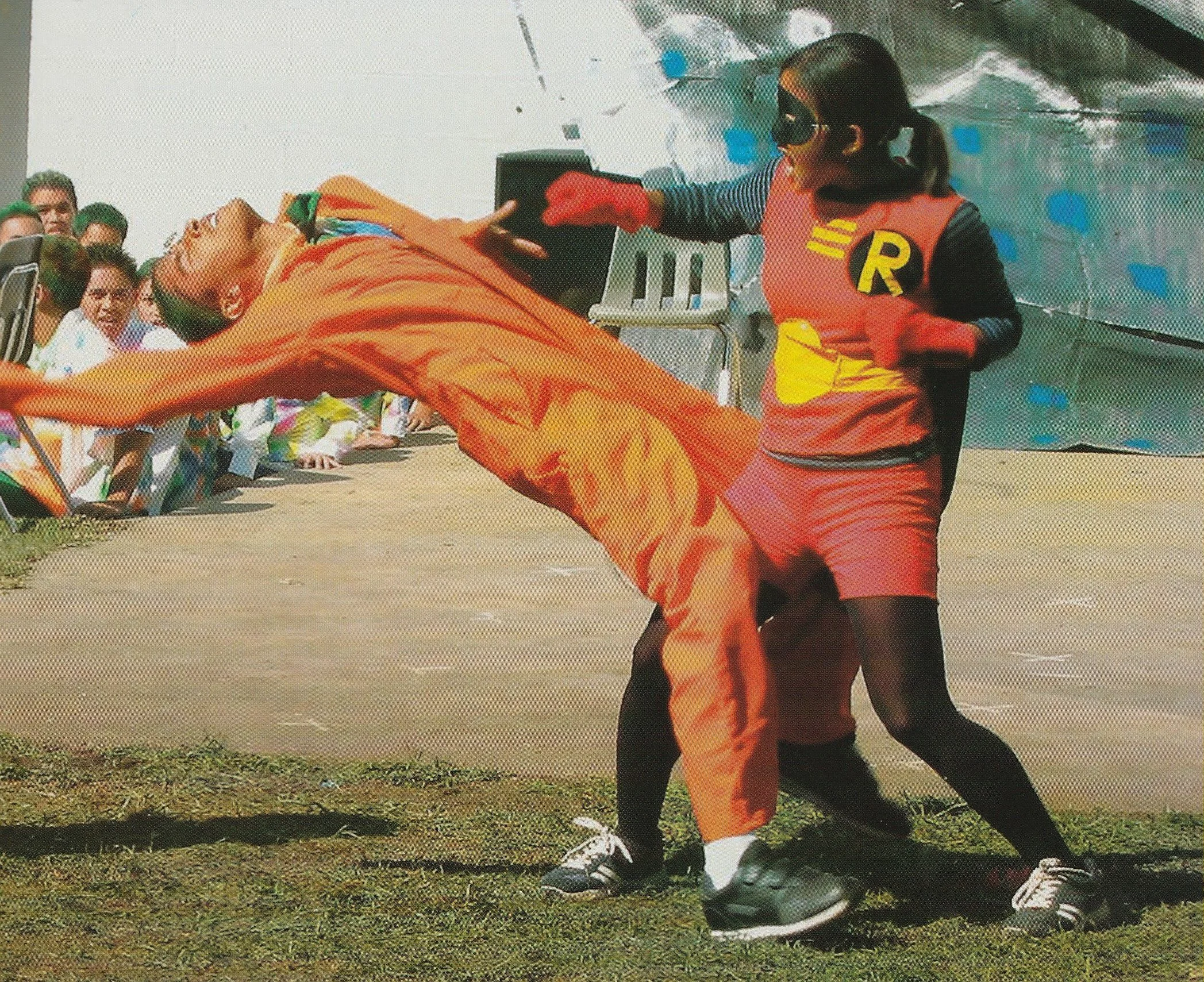 A person dressed as Batgirl is grabbing another individual dressed in orange costume from the front during a staged outdoor event, with a crowd of onlookers watching.