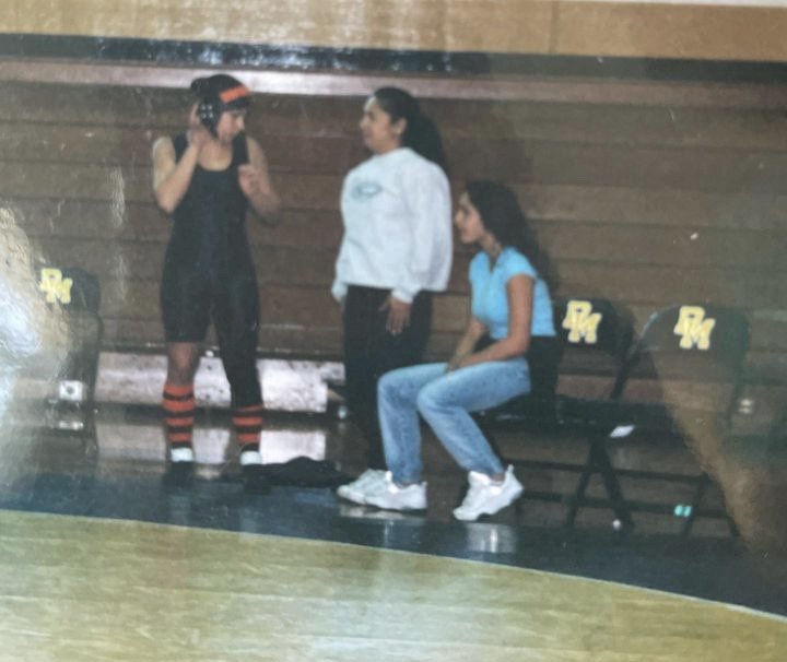 Three young women in a gym or sports practice area. One woman standing in athletic gear, black with red striped socks, and the other two sitting on chairs wearing casual workout clothes. There are black chairs with yellow letters on the backrests, and a wooden wall in the background.