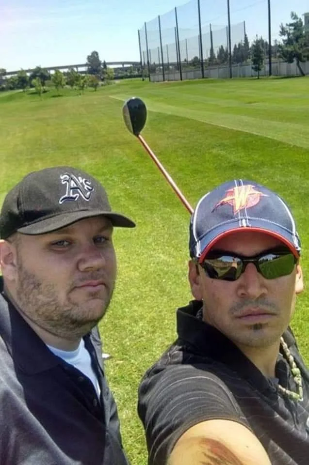 Two men taking a selfie on a golf course with a golf club positioned behind their heads to resemble bunny ears.