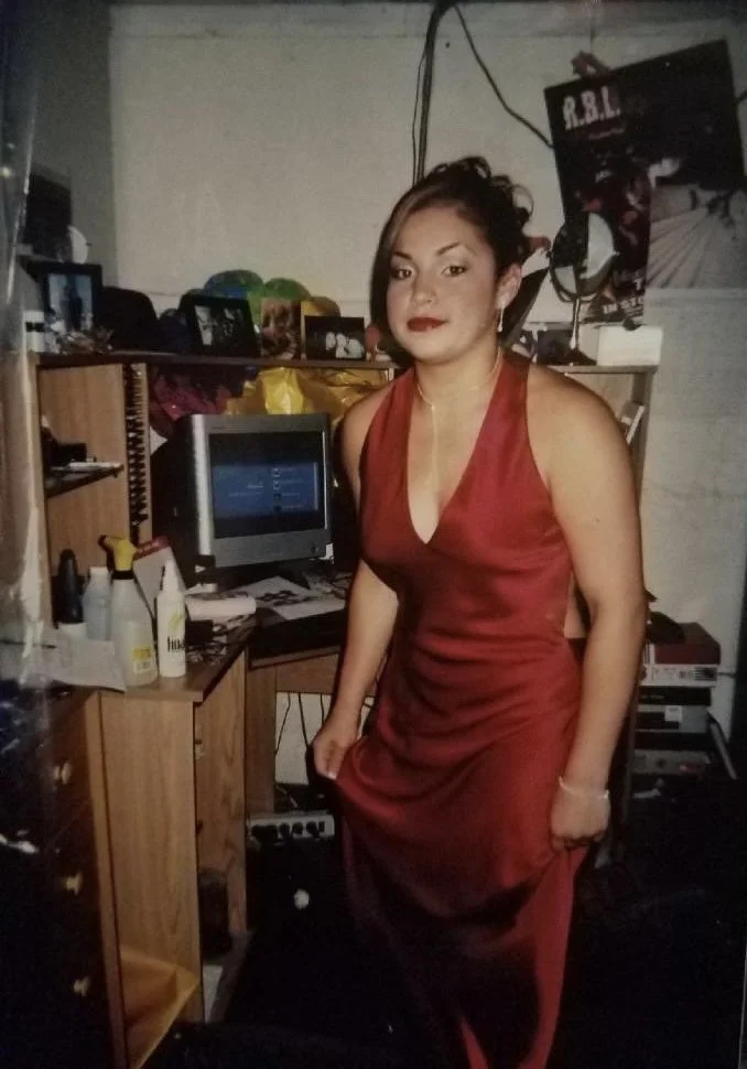 Woman in a red satin dress standing in a cluttered room with a computer, books, and miscellaneous items on shelves.