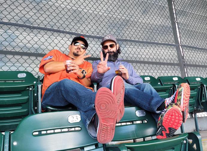 Two men sitting on green stadium seats behind a chain-link fence, wearing casual clothes, sunglasses, and hats, holding drinks, with one making a peace sign, and both showing their shoe soles toward the camera.