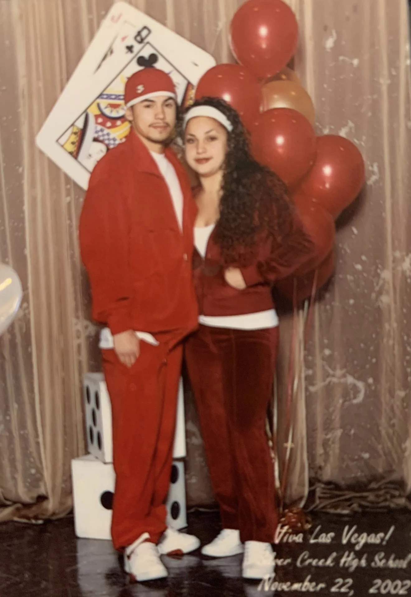 A young man and woman dressed in red, standing close together in front of a balloon and playing card decoration, at an event celebrating Viva Las Vegas, November 22, 2002.