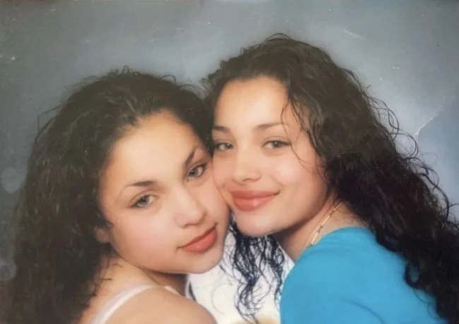 Two young women with curly hair smiling and leaning close together, posing for a photo against a gray background.