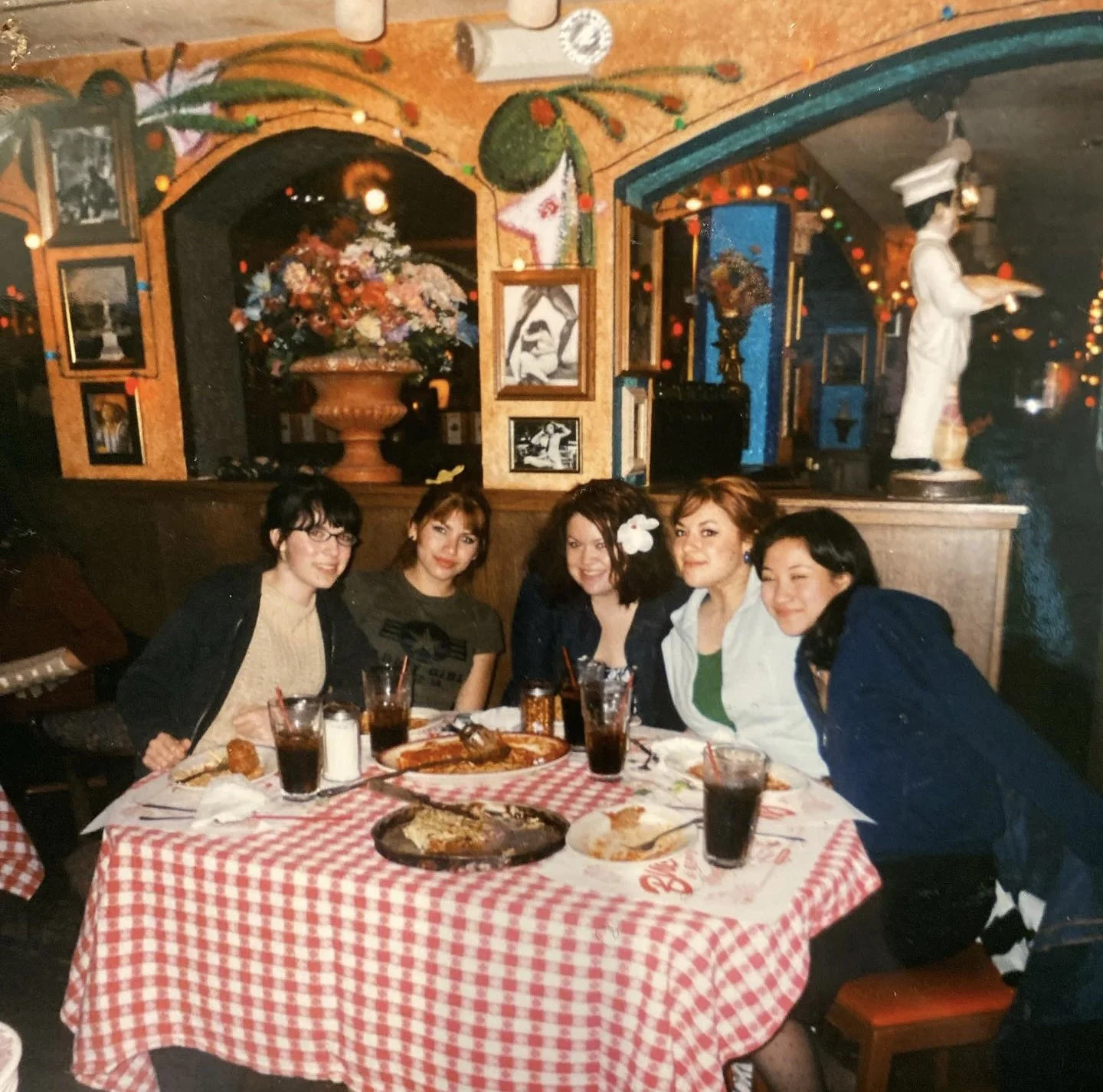 Five women sitting around a table with food and drinks in a cozy restaurant decorated with colorful artwork, flowers, and a statue in the background.