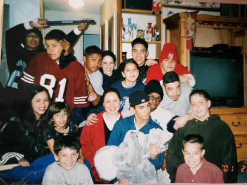Group of children and teenagers gathered together in a living room, some sitting on the floor and others standing behind, smiling and posing for a photo.