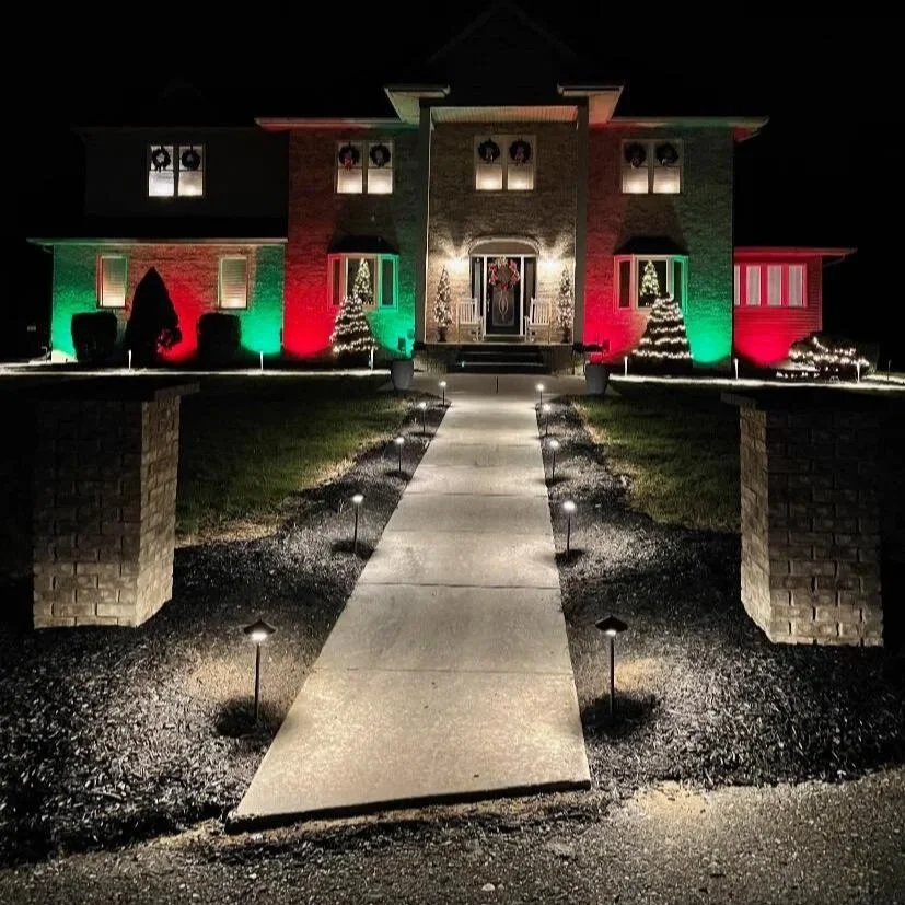 A house decorated for Christmas with red and green lights illuminating the exterior, Christmas trees, and festive decorations, viewed at night.
