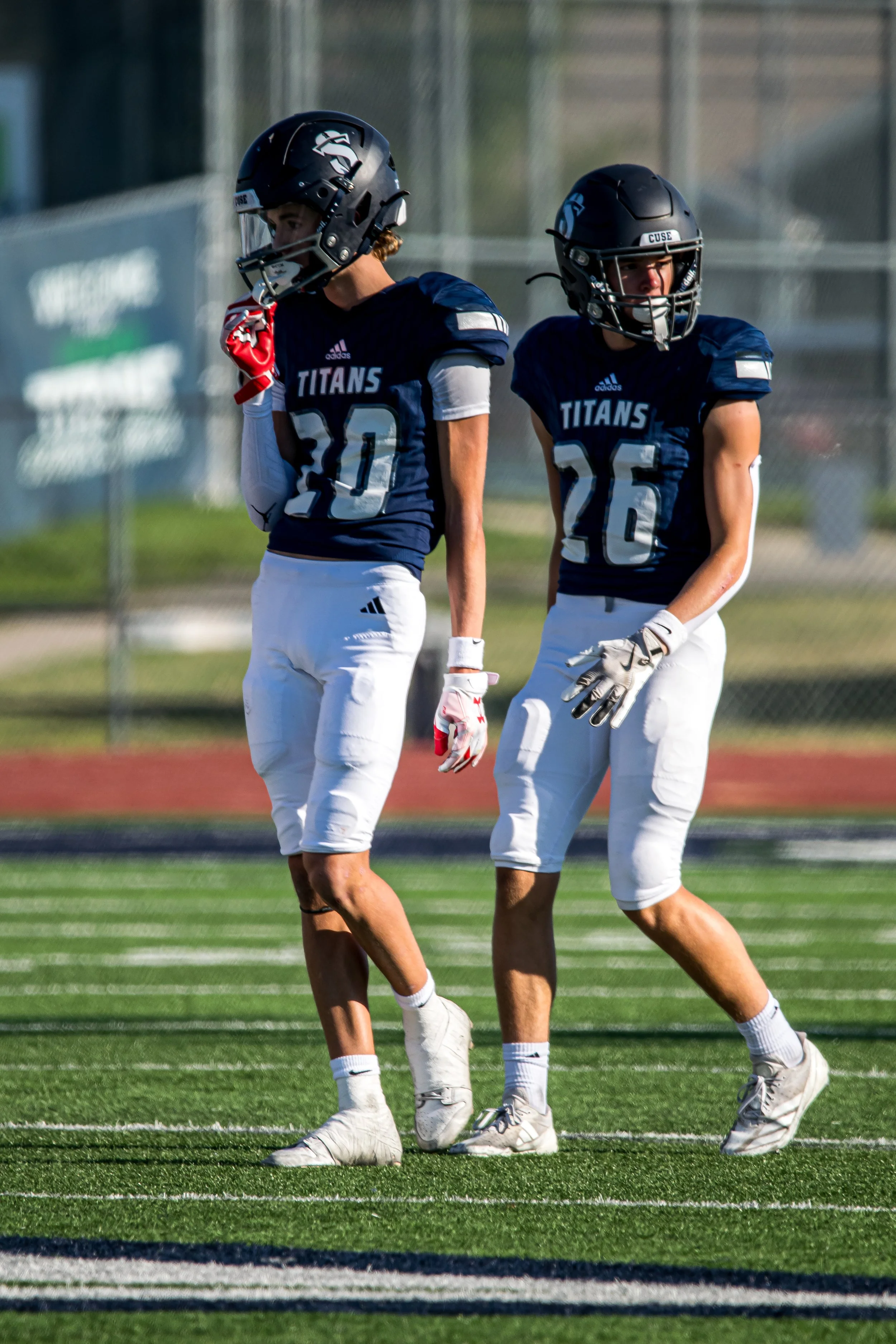Two football players in navy blue and white uniforms, wearing helmets and gloves, standing on a football field during practice.