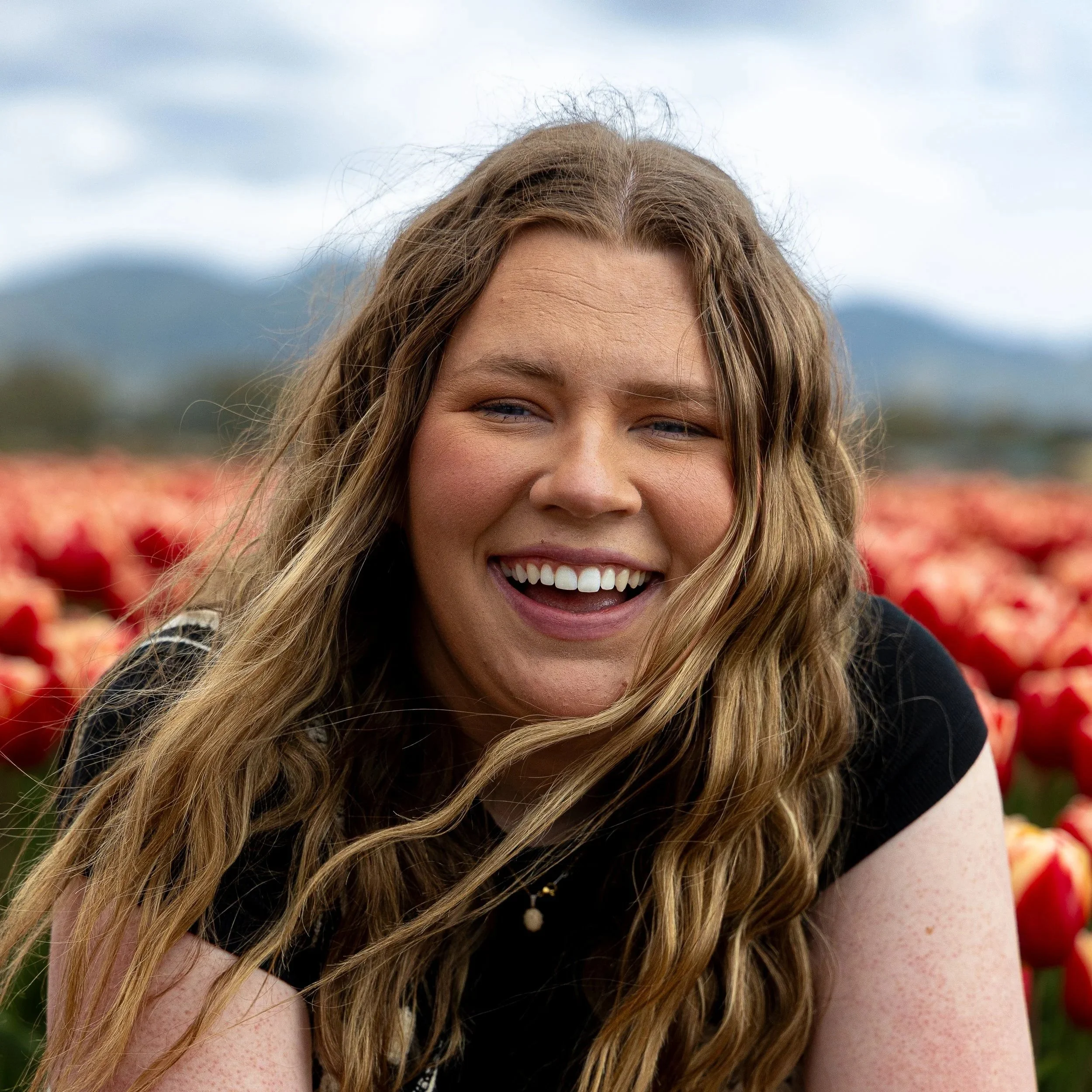 A woman with long wavy hair smiling outdoors in a field of red tulips with mountains in the background.