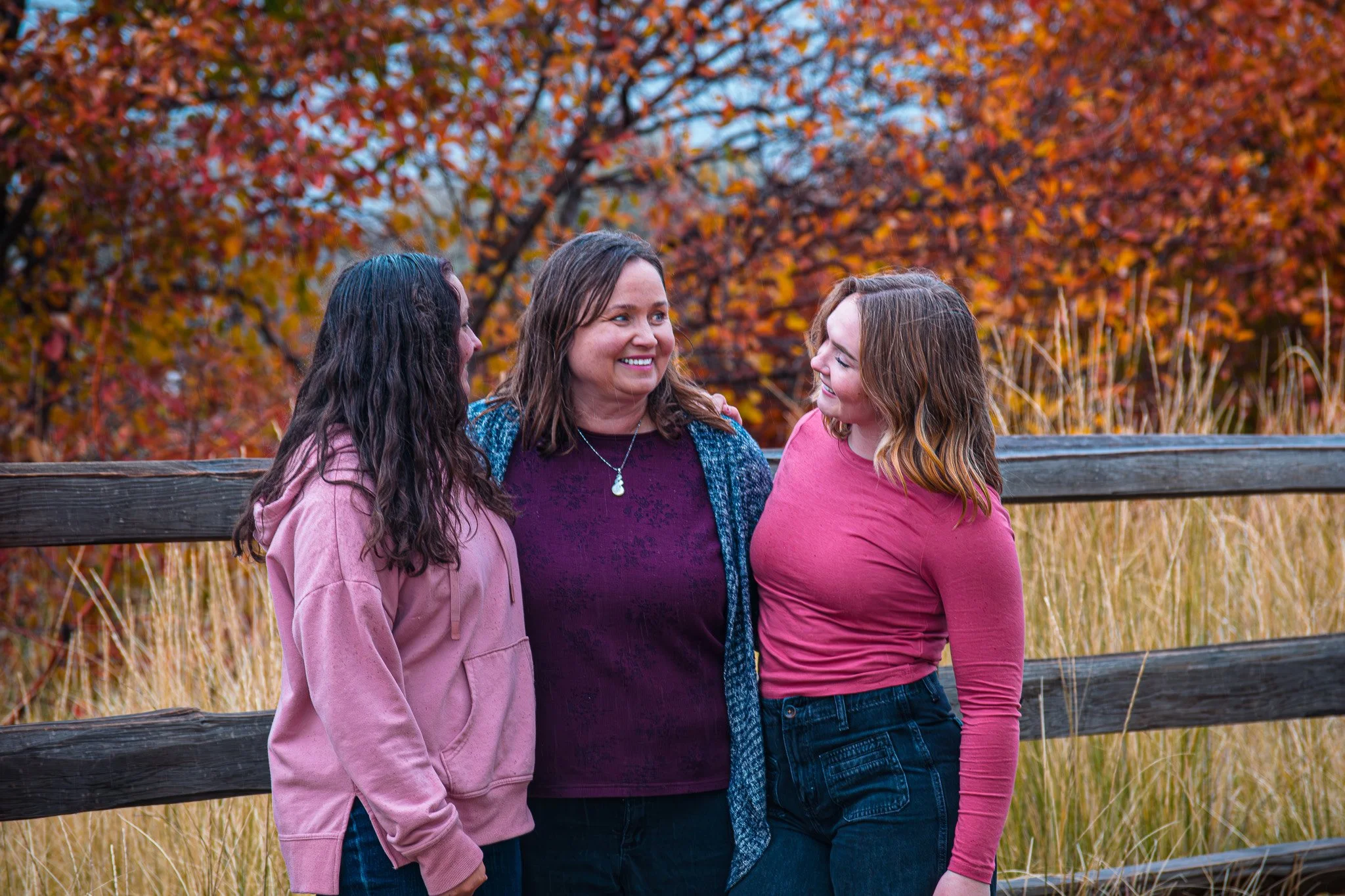Three women standing outdoors in front of autumn-colored trees and grass, smiling and talking.