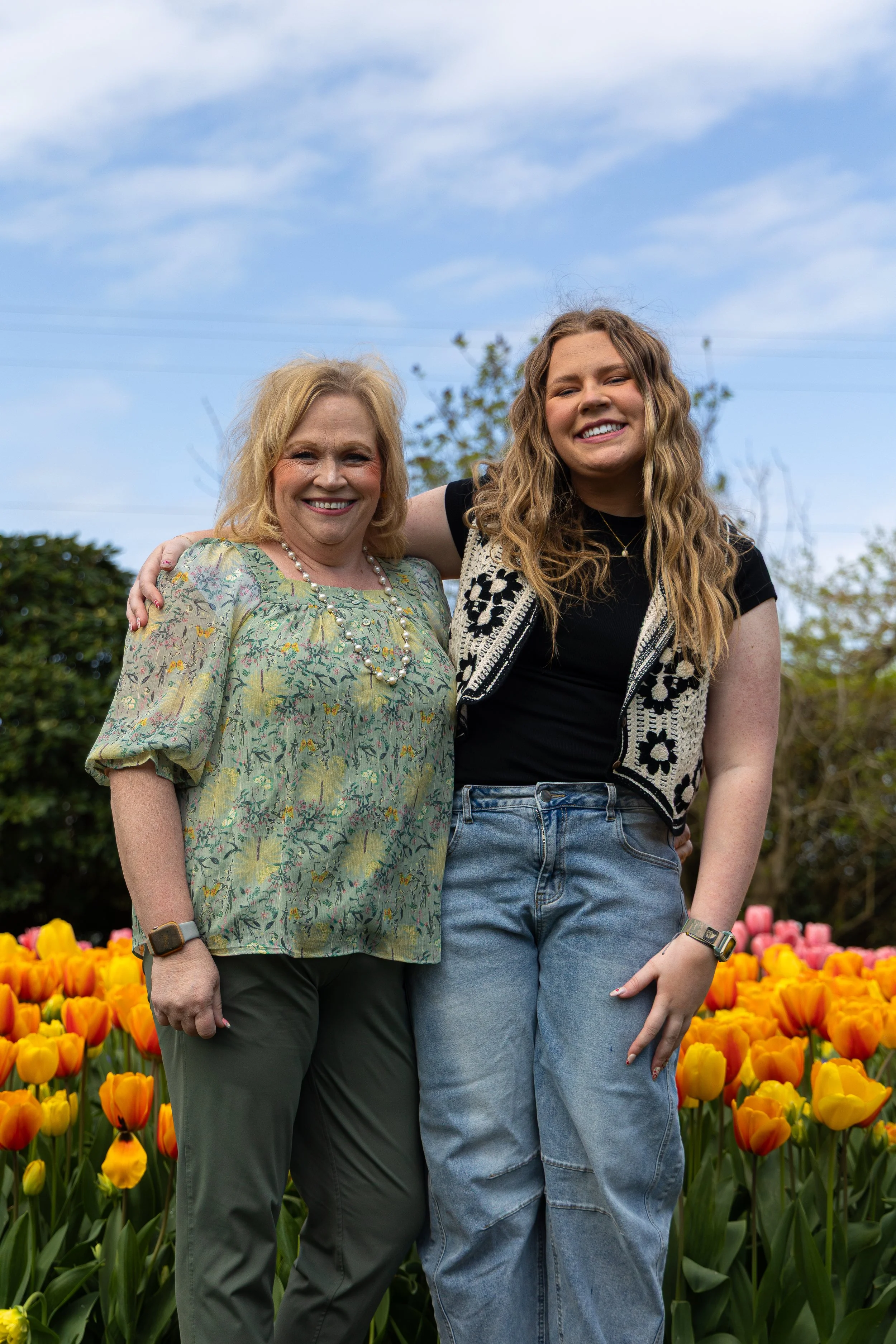 Two women standing close together in a garden with tulips, smiling at the camera. One woman has blonde hair, wearing a floral blouse and dark pants, while the other has long, wavy hair, wearing a black shirt with a patterned vest and jeans.