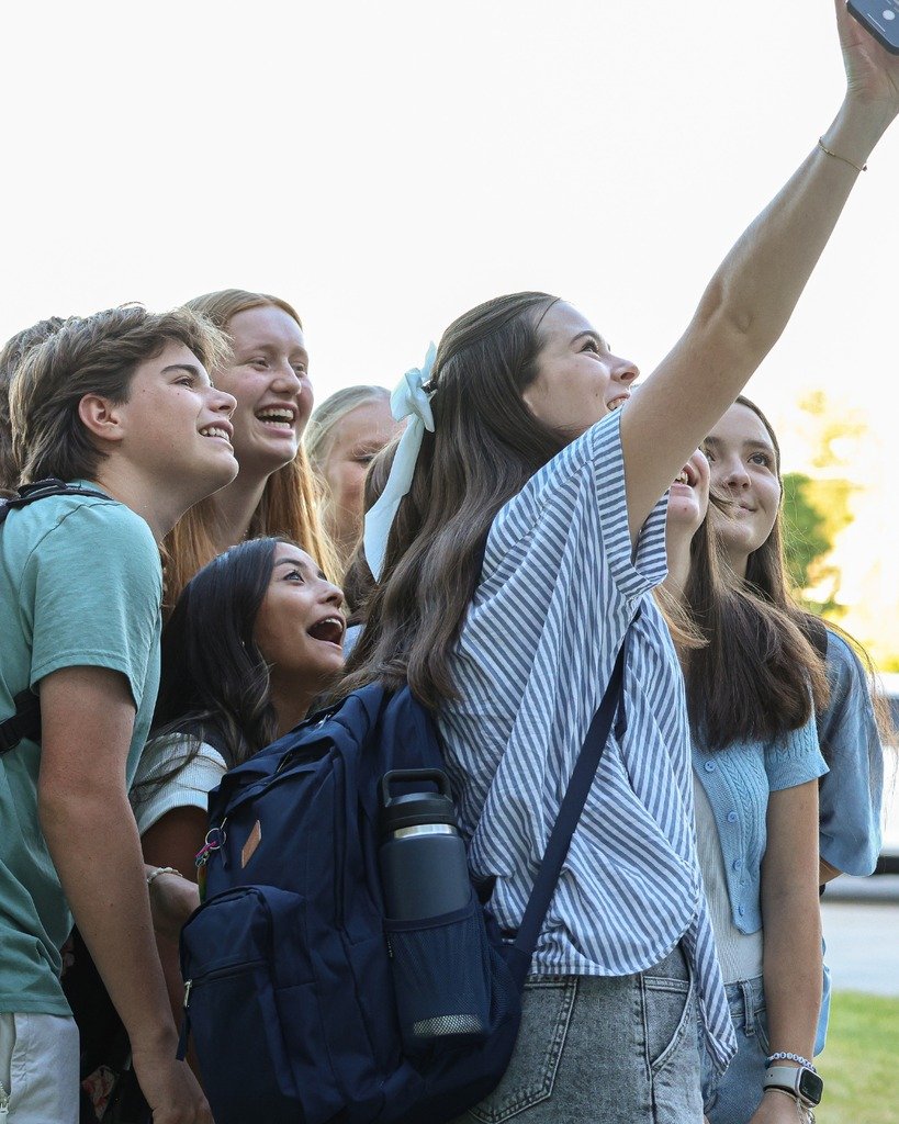 Group of six young girls taking a selfie outdoors on a sunny day, smiling and enjoying each other's company.