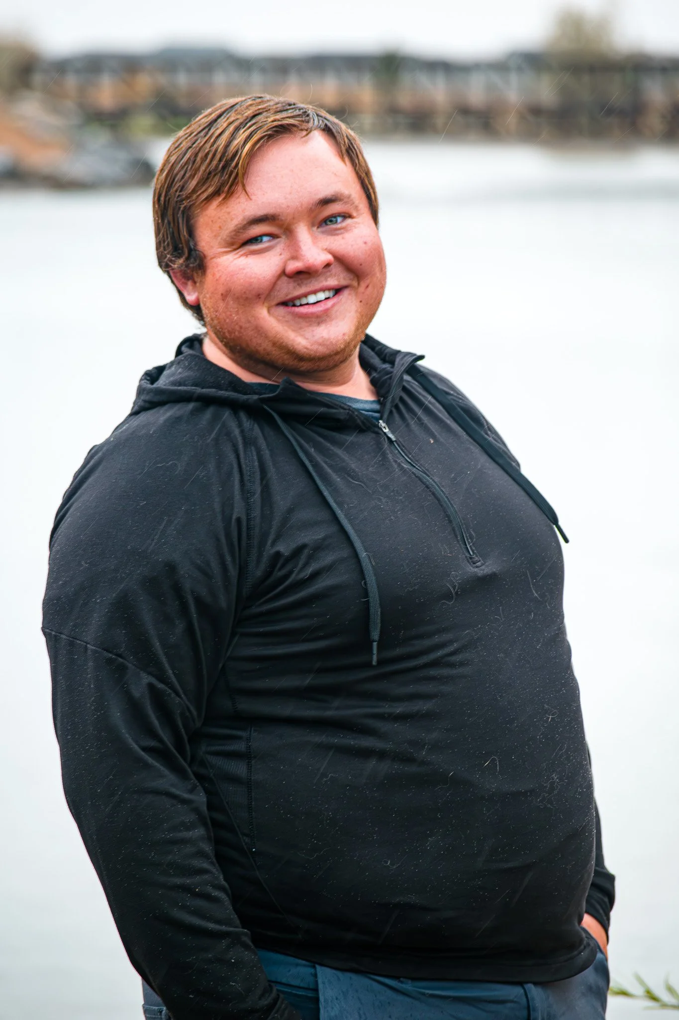A smiling young man with light brown hair and blue eyes stands outdoors near a body of water, wearing a black hooded jacket.