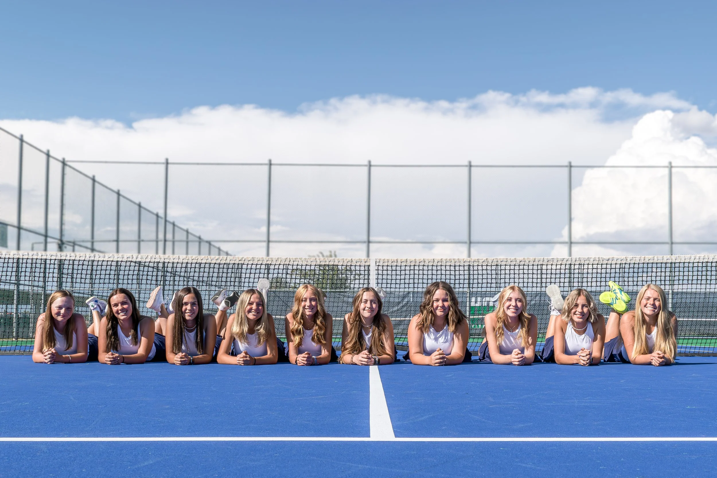 Group of ten young women in tennis uniforms laying on a blue tennis court in a line, smiling with their hands clasped, with a tennis net and sky in the background.