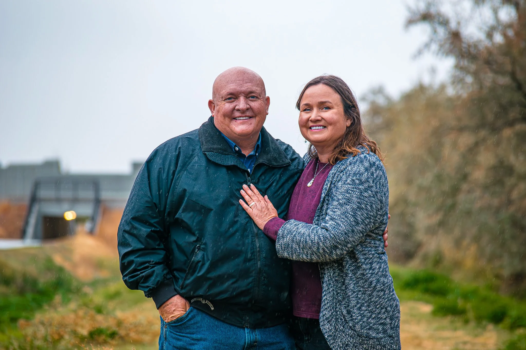 A couple standing outdoors in the rain, smiling at the camera, with trees and blurred background.