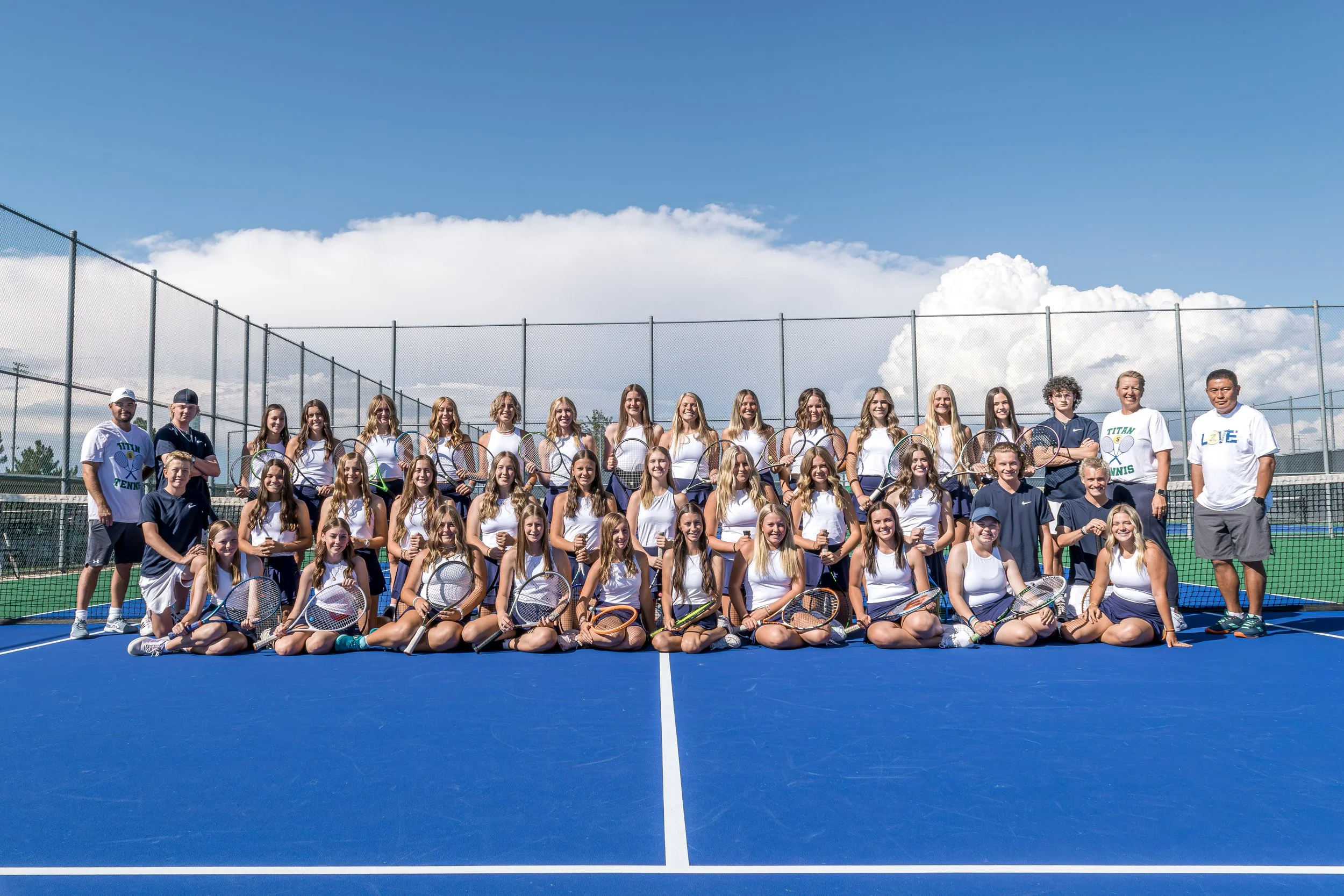Group of young female tennis players and coaches on an outdoor blue tennis court, with a chain-link fence and cloudy sky in the background.
