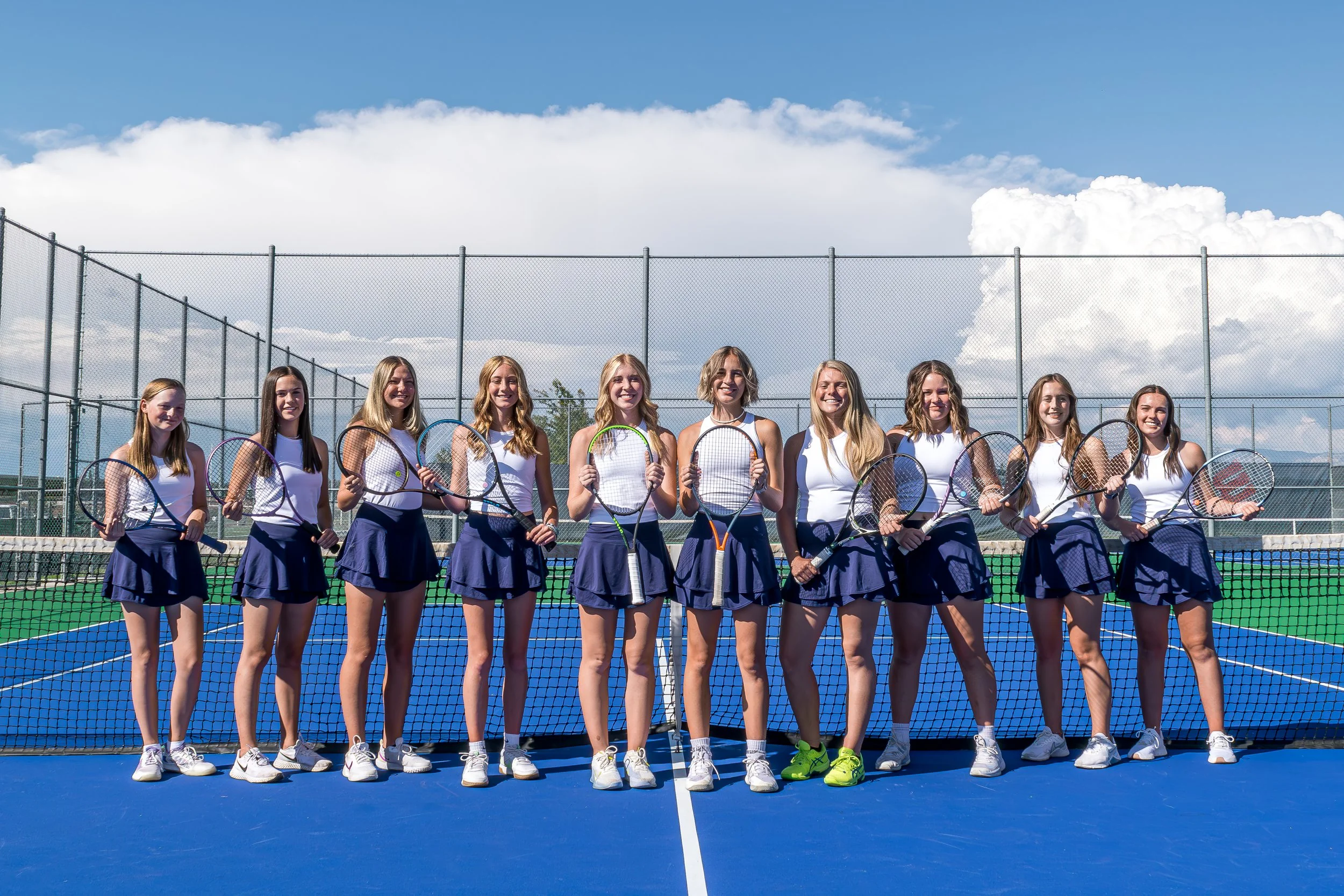 A group of ten young women in tennis outfits standing in a line on a tennis court, holding tennis rackets, with a fence and blue sky with clouds in the background.