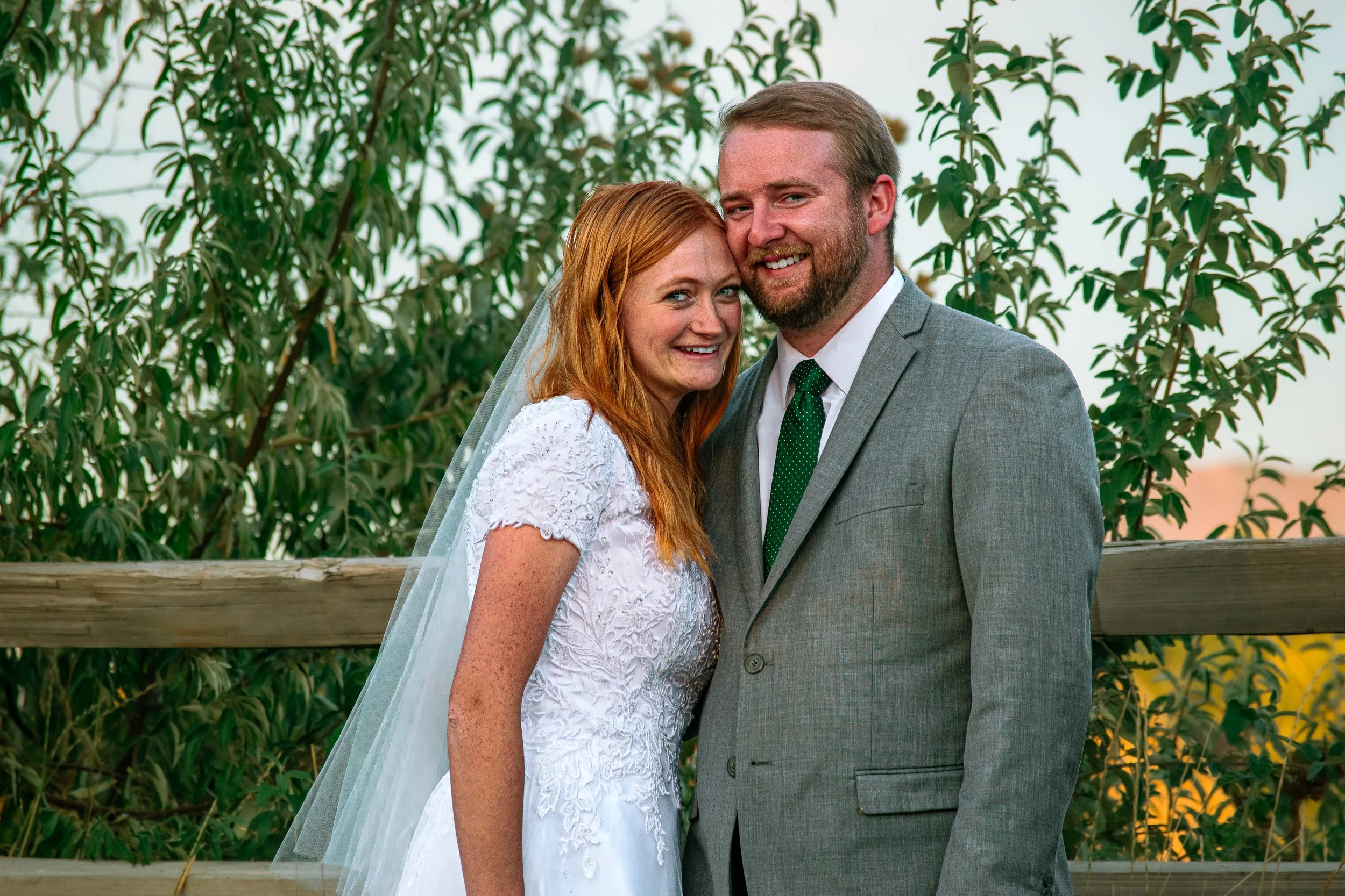 A bride and groom stand closely together outdoors, smiling at the camera, with greenery and a wooden fence in the background.