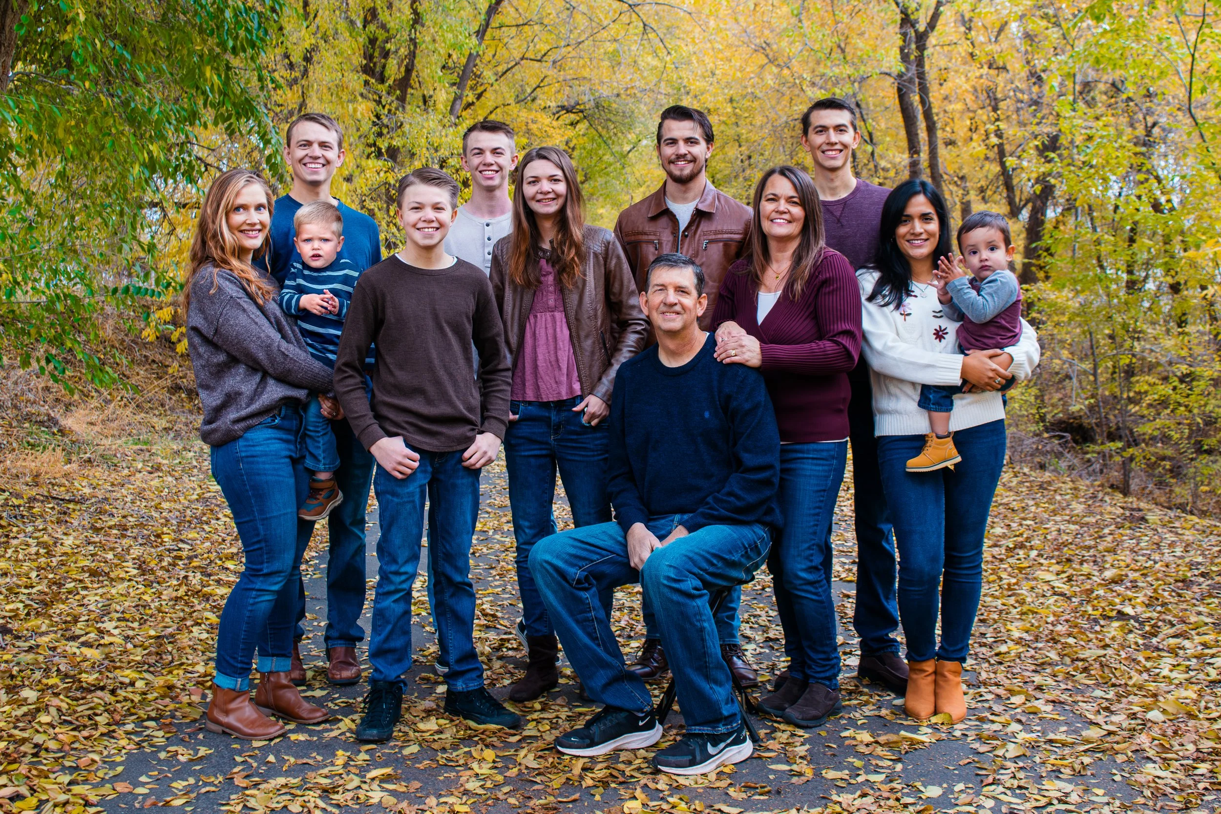 Family of thirteen people outdoors during autumn, surrounded by yellow and green trees, standing on a leaf-covered path, smiling at the camera.