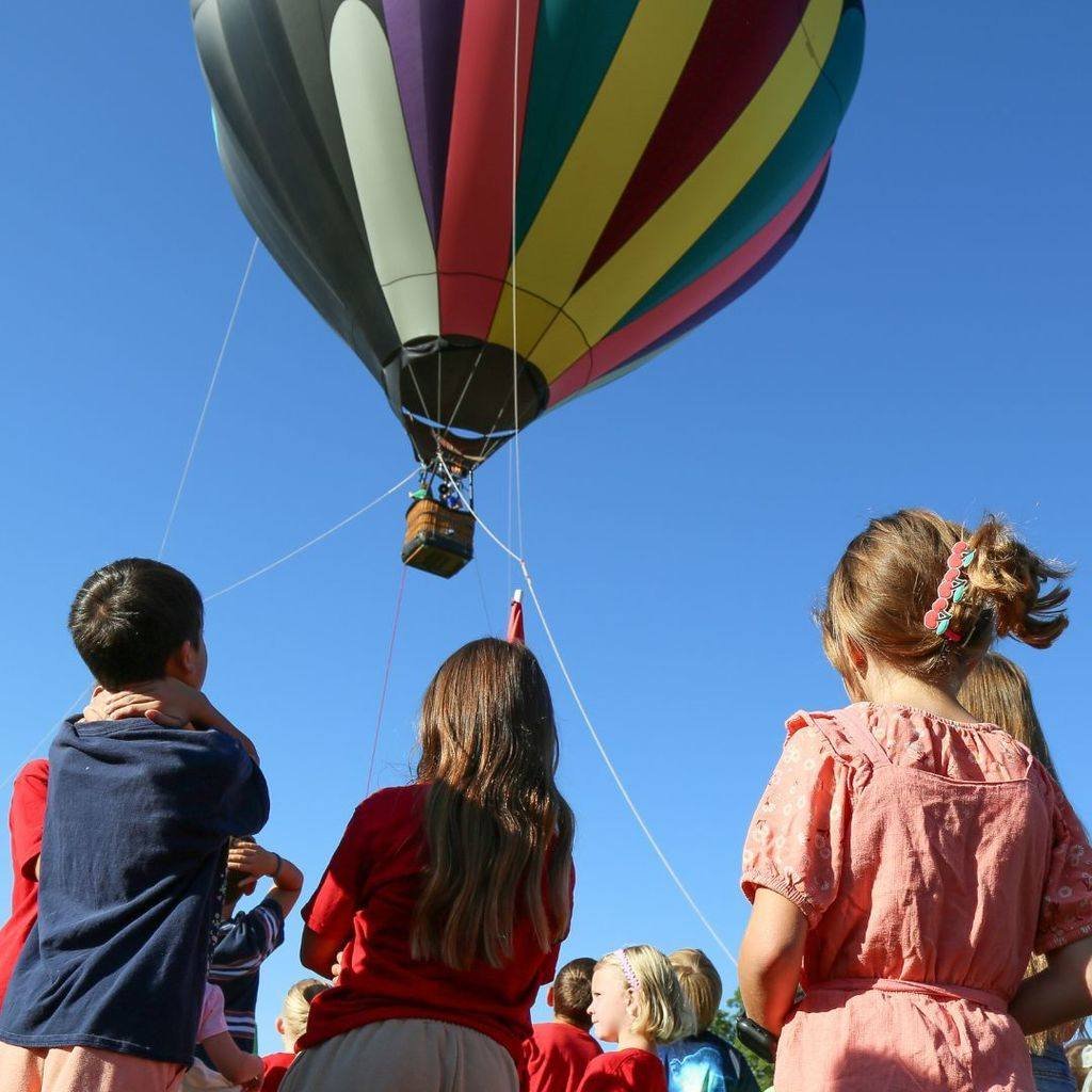 Children watching a colorful hot air balloon fly in a clear blue sky.