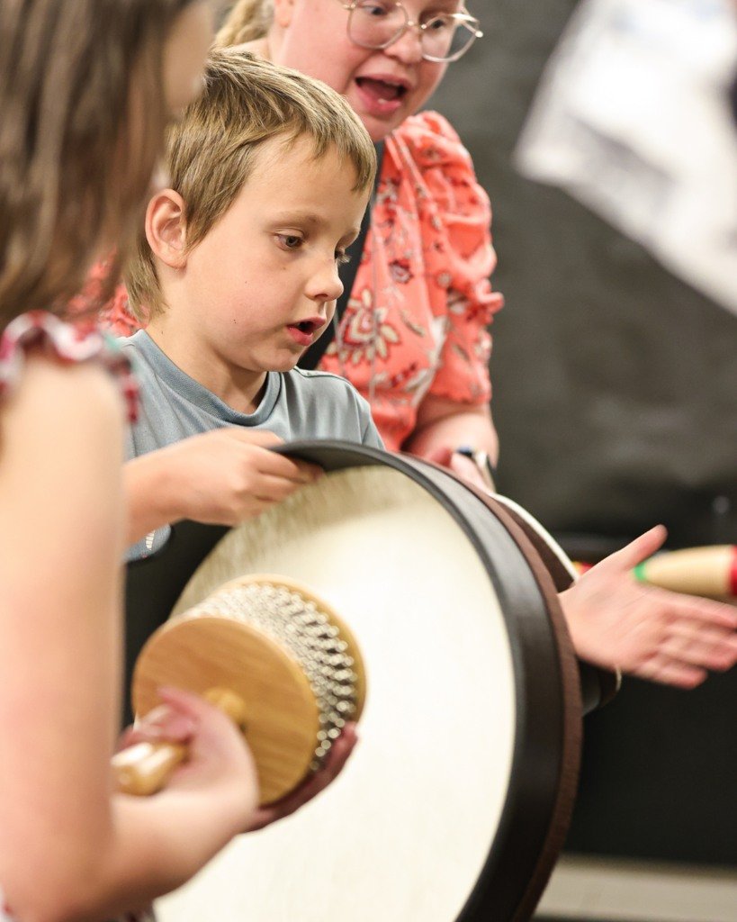 A young boy looks at a large drum being played by an adult, with another adult holding a mallet, in a classroom or indoor setting.