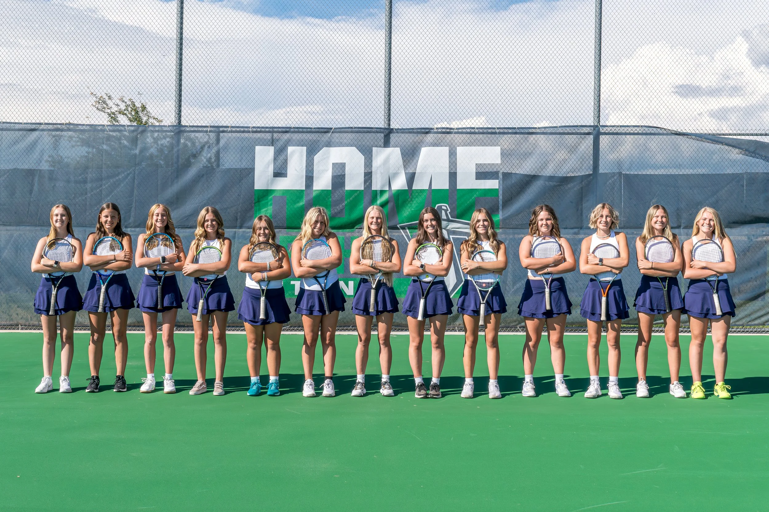 Group of young female tennis players standing on a tennis court with arms crossed, holding tennis rackets, in front of a backdrop with the words 'HOME TEAM' and a tennis logo.