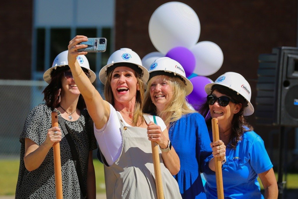 Four women wearing construction helmets and holding shovels, smiling and taking a selfie at an outdoor event with balloons in the background.