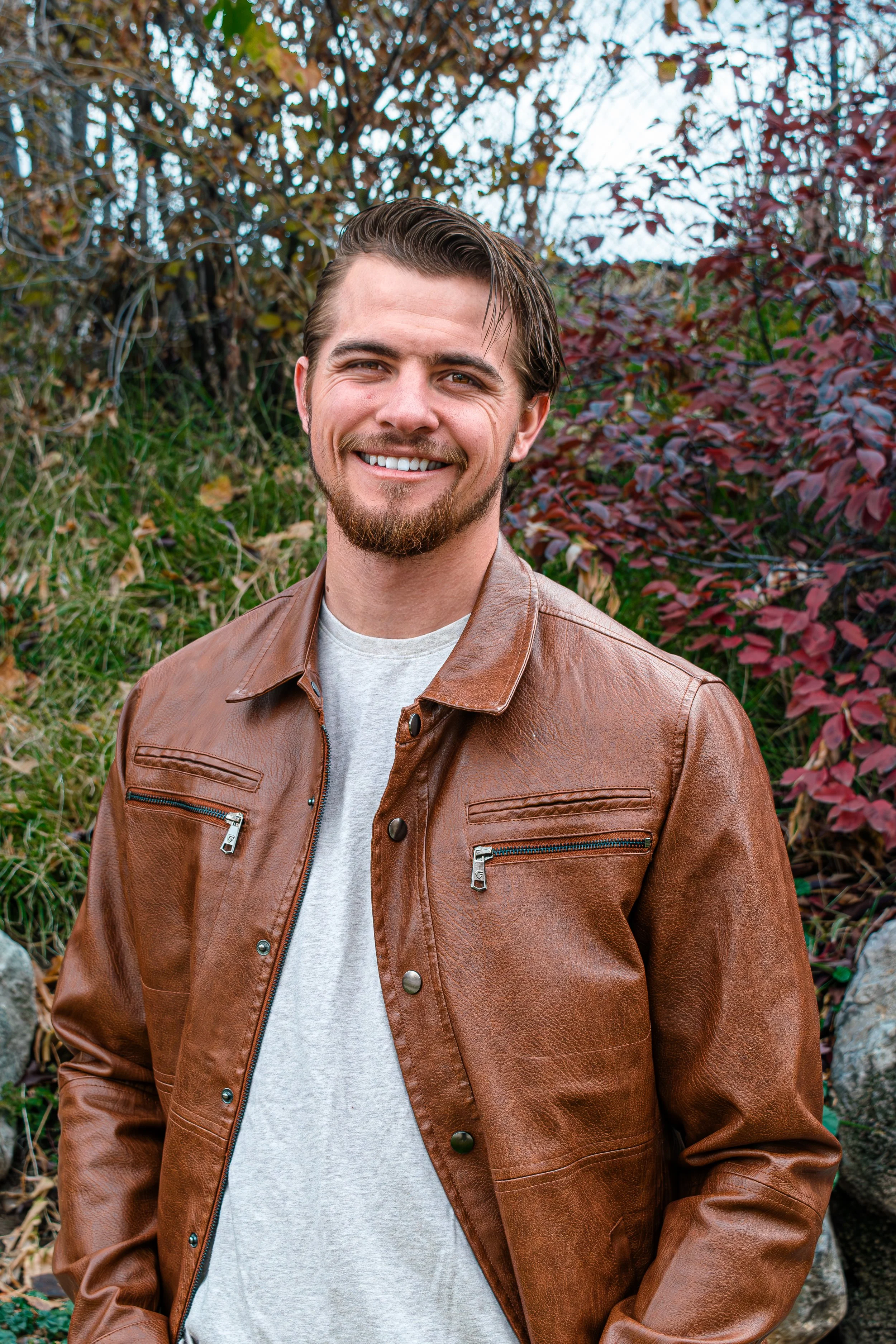 A smiling man with a beard and short dark hair, wearing a brown leather jacket over a light gray shirt, standing outdoors with trees and foliage in the background.