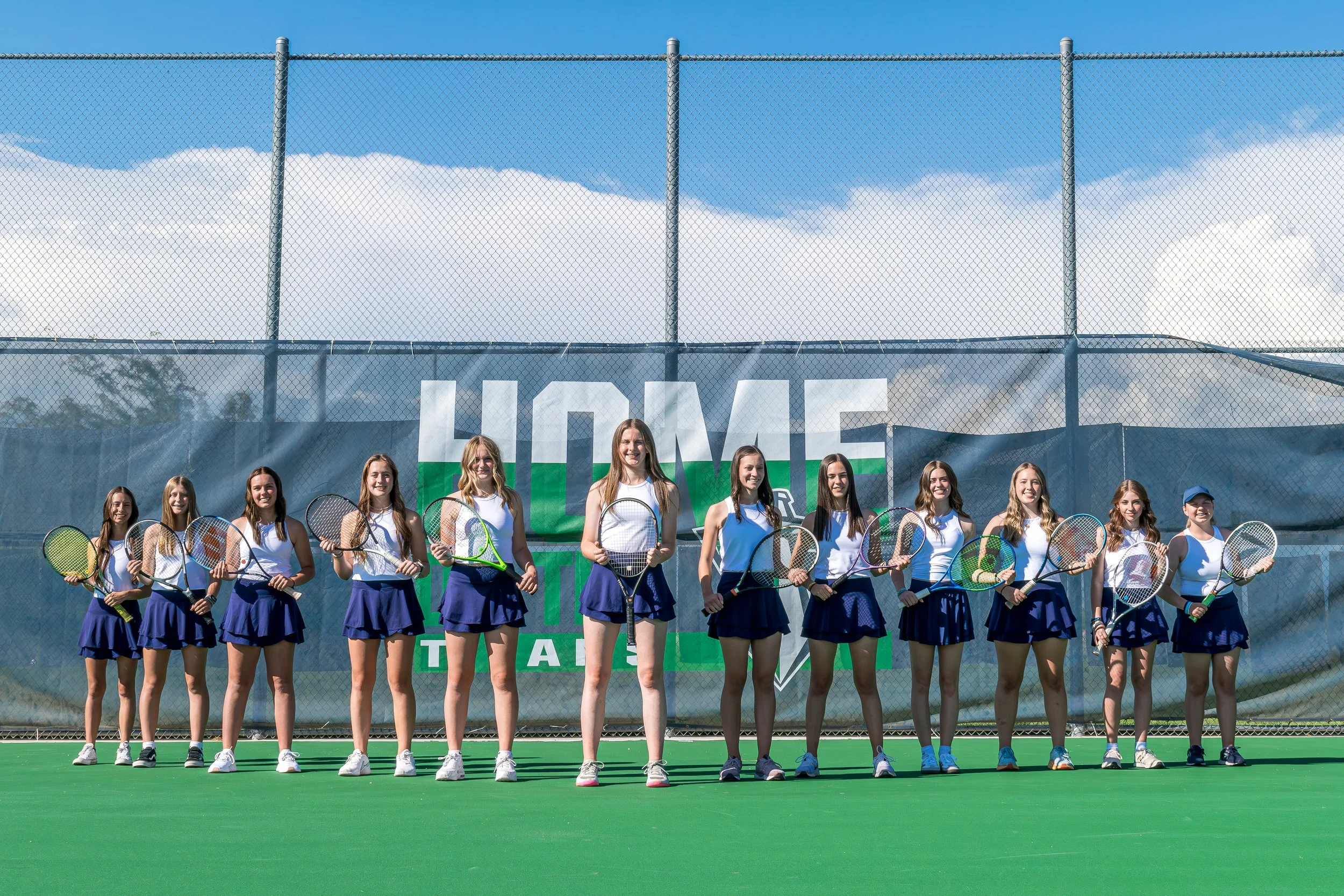 A group of young women tennis players in uniform standing on a tennis court, holding tennis rackets, in front of a large sign with the word 'HOME' on it.
