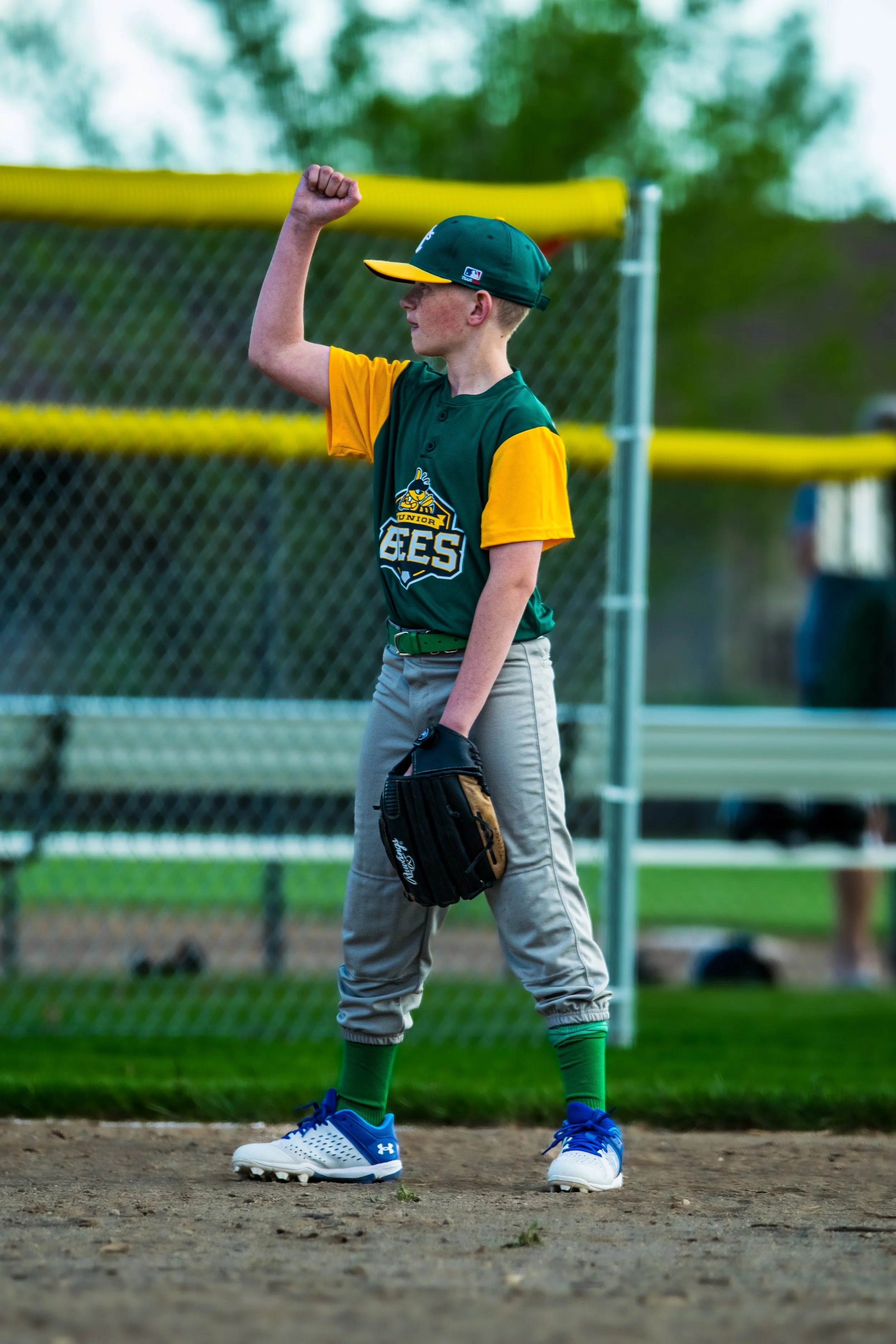 A young baseball player in a green and yellow uniform, standing on a baseball field with a glove, raising his fist in a gesture of confidence or celebration.